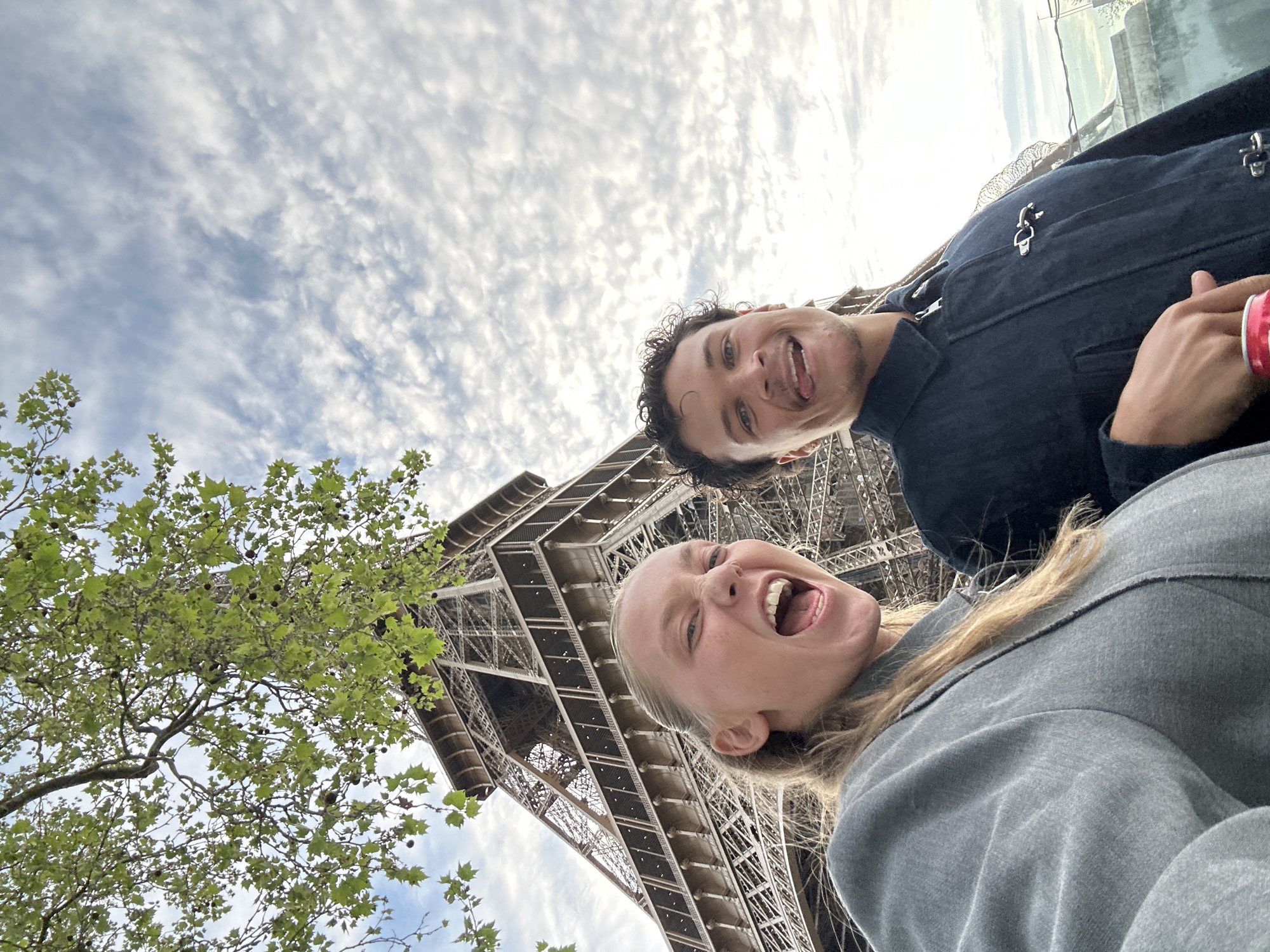 Two people are posed in front of the Eiffel Tower in Paris, France. A woman with long blonde hair pulled back, and wearing a grey top, is on the bottom. A man with curly dark hair and wearing a dark-colored jacket is above her. Both have their mouths open and are smiling. A tree with green leaves is in the lower left corner. The background consists of the Eiffel Tower, a cloudy sky and rooftops of adjacent buildings.