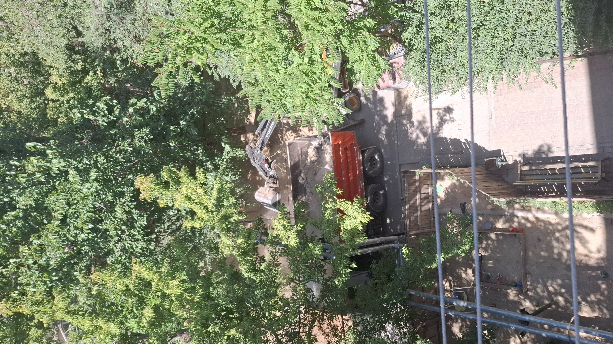 An aerial view captures a construction scene in Shymkent, Kazakhstan. A red dump truck is being loaded with dirt and debris by a black Volvo excavator. The excavator's bucket is positioned over the truck's bed, scooping a load of light-colored material. The truck has dual rear wheels and appears to be parked on a paved surface. Lush green trees surround the work area, casting shadows on the ground. In the foreground, a fenced-off area contains a sandpit with various toys scattered within it, suggesting it might be a children's play area. Wooden fencing and benches are also visible. The overall scene depicts an ongoing construction or demolition project occurring in an urban environment.