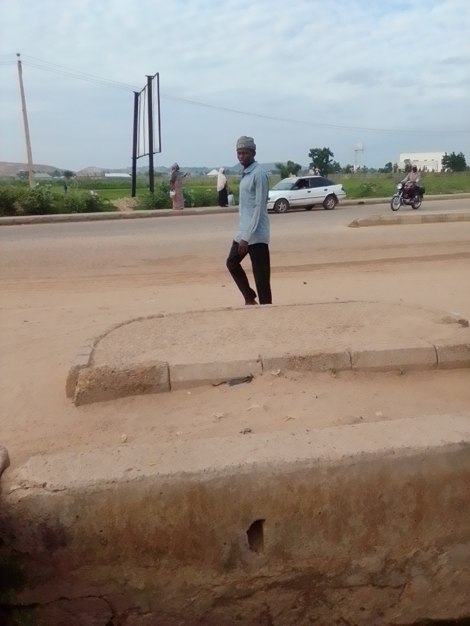 In Kwadon, Nigeria, a young man in a light blue long-sleeved shirt and dark pants walks across a dusty, light-colored road. He is wearing a gray cap. To his left, a white sedan is parked. In the background, a motorcycle with a rider is visible on the road. Further back, there are green fields and some buildings, including what appears to be a water tower and a large white structure. The sky is overcast. A billboard structure is also present in the distance. Several people are seen standing and walking along the roadside in the middle ground. The foreground consists of a concrete structure with rough edges.
