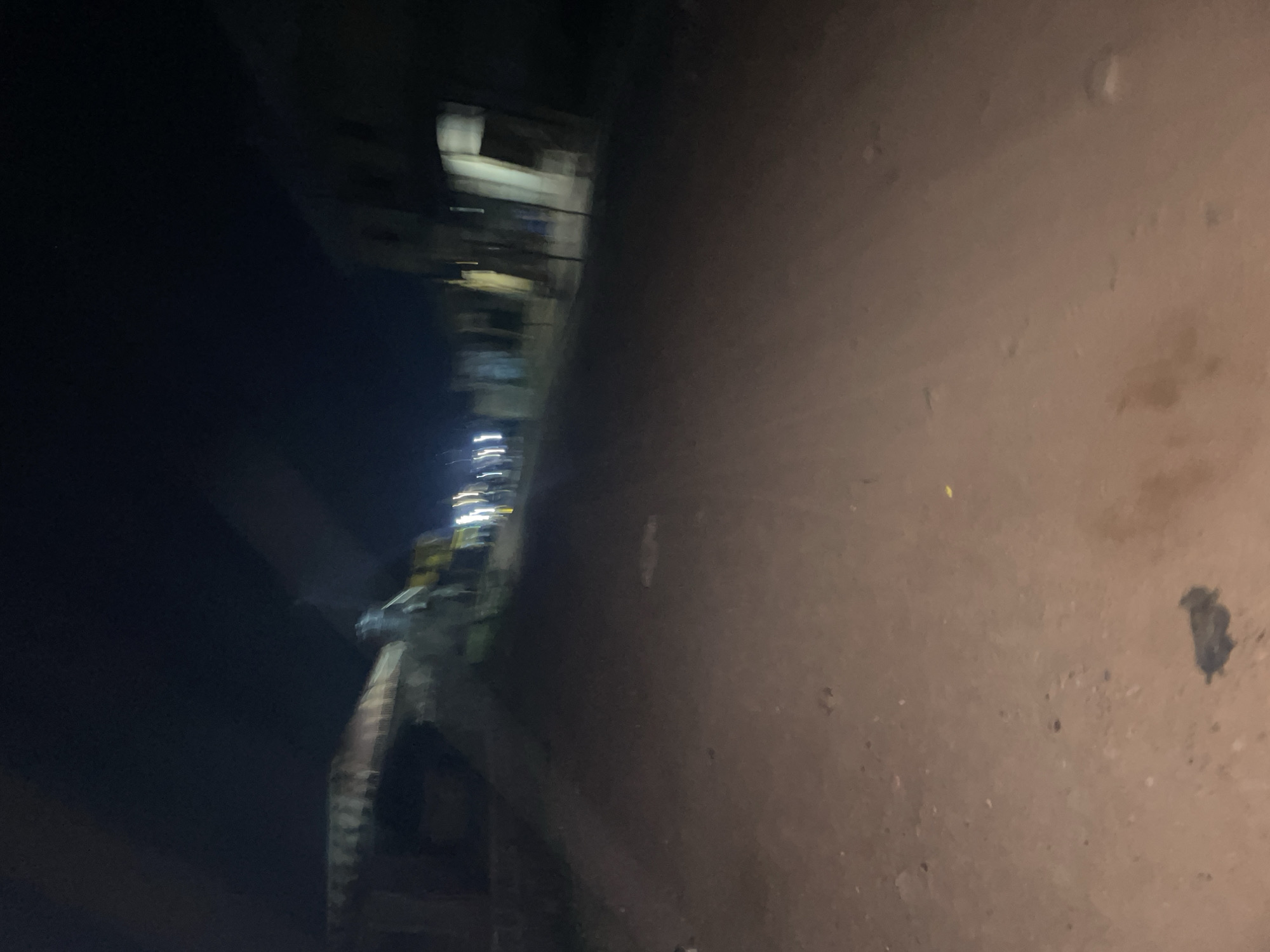 The image depicts a dimly lit dirt road at night in Lokoja, Nigeria. The road appears to be unpaved and is surrounded by buildings on either side, some of which show signs of habitation with lights on. There are several blurred lights visible in the distance, possibly from vehicles or street lamps, contributing to the overall low-light conditions. The environment suggests a residential or commercial area within the city, likely with limited infrastructure given the dirt road. The low resolution and motion blur make it difficult to identify specific individuals or detailed objects.