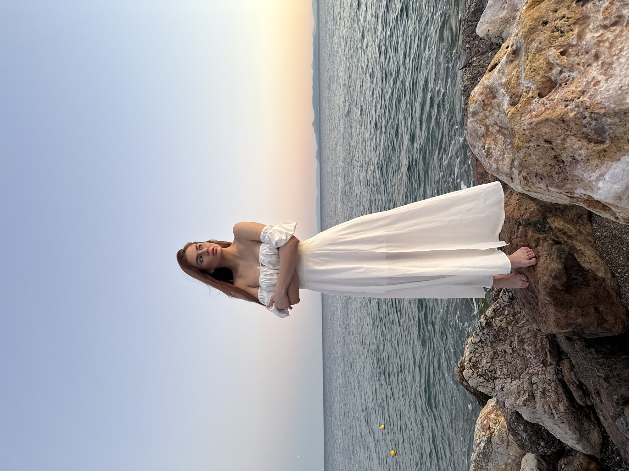 A young woman with long, auburn hair stands on a rocky shore in Paleo Faliro, Greece. She is wearing a flowing white off-the-shoulder dress and has her arms crossed. The sea is calm and a distant mountain range is visible on the horizon. The sky is a clear blue, transitioning to a soft orange and pink at the horizon, indicating either sunrise or sunset. Yellow buoys are visible in the water. The rocks beneath her are varied in size and color, with some showing signs of lichen growth.
