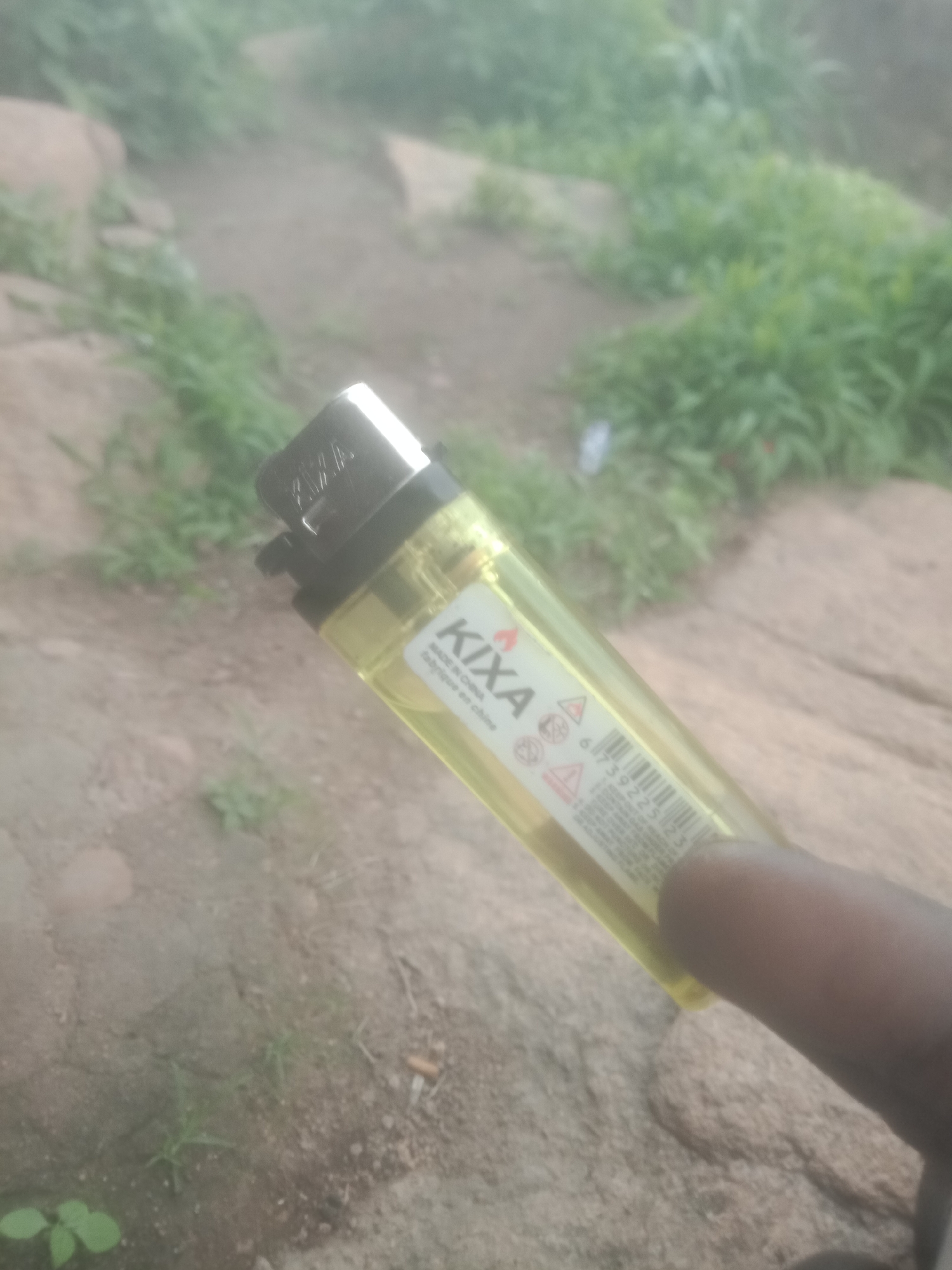 A yellow disposable lighter is held in a person's right hand. The lighter has a black cap and a label with the brand name "KIXA" printed in white. Below the brand name, there is a barcode and some text in a smaller font. The background is out of focus, showing rocks and vegetation. The scene appears to be outdoors in a natural setting.