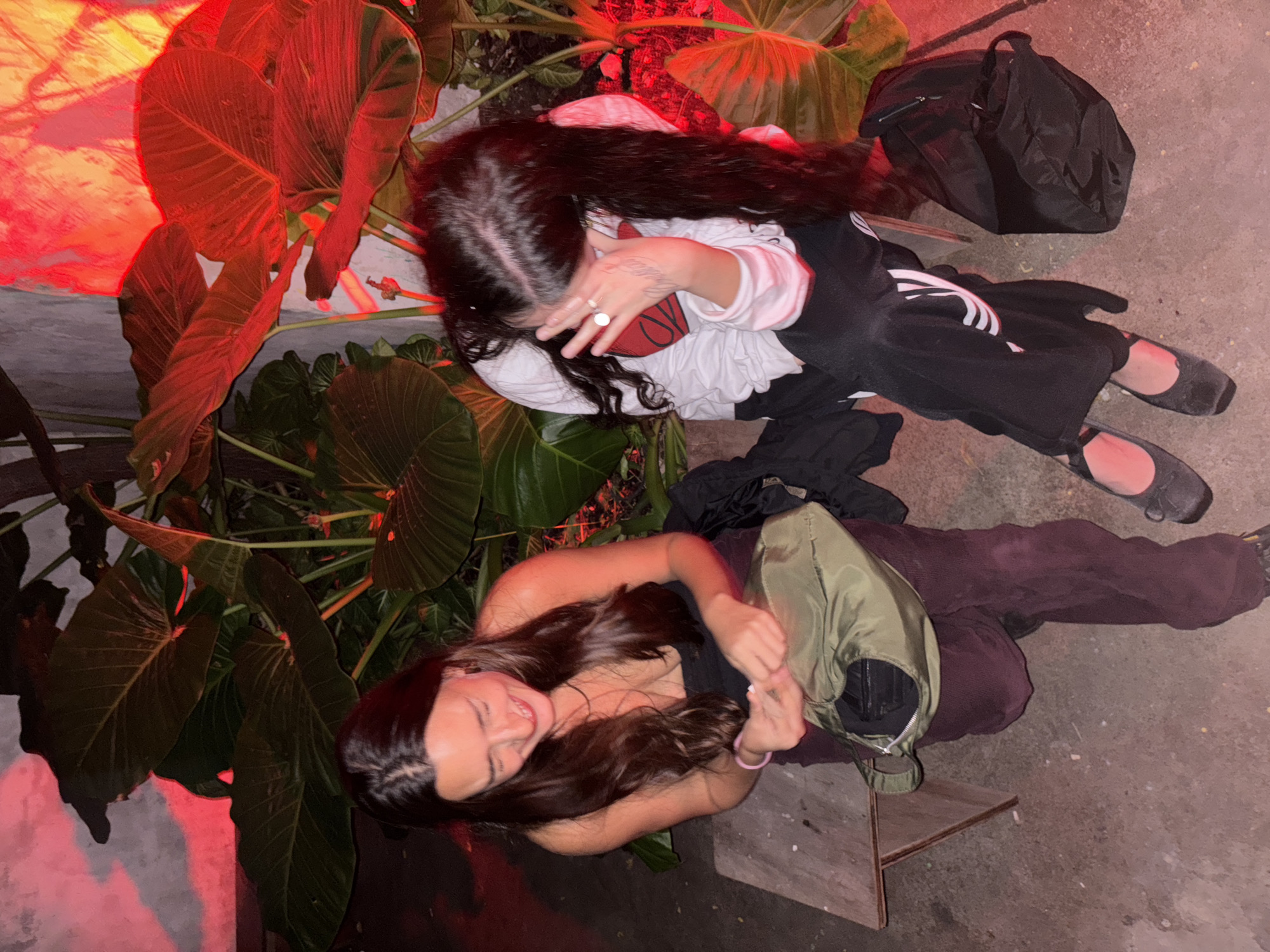 Two young adult women are sitting on the ground outdoors. One woman is sitting on a small, wooden bench, wearing a black top and burgundy pants, holding an olive green bag. She has long, brown hair and is smiling. The second woman is sitting on the ground, wearing a white long-sleeved shirt with a graphic design, black track pants with white stripes, and black flats. She has long, black hair and is covering her face with her hand. Behind them are large green plants and a red light. A black backpack is on the ground. The location appears to be outdoors, possibly a patio or urban area. The image was taken at night. There is no explicit information that can determine the location in the city of Buenos Aires, Argentina.