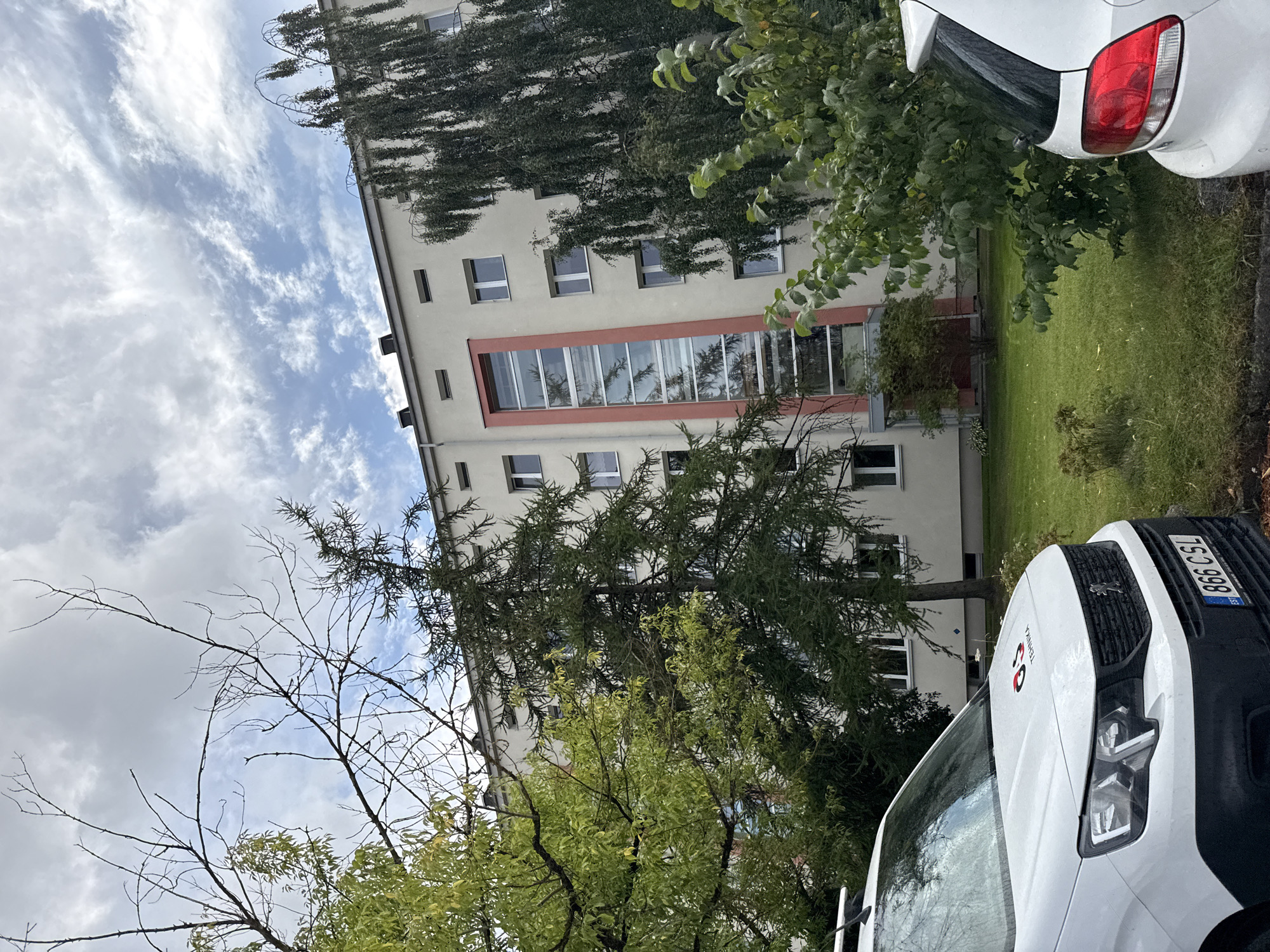 The image depicts a white van parked on a grassy area in front of a light-colored apartment building. The van has a logo on its hood that reads "OS TEHNIKA" and an Estonian license plate with the characters "866 CSL". The building features multiple windows and a vertical, red-trimmed glass section. Trees with green foliage and bare branches are visible surrounding the building and the van. The sky is partly cloudy. The scene appears to be a residential or commercial area in Tallinn, Estonia, based on the license plate and building style. There are no people visible in the image.