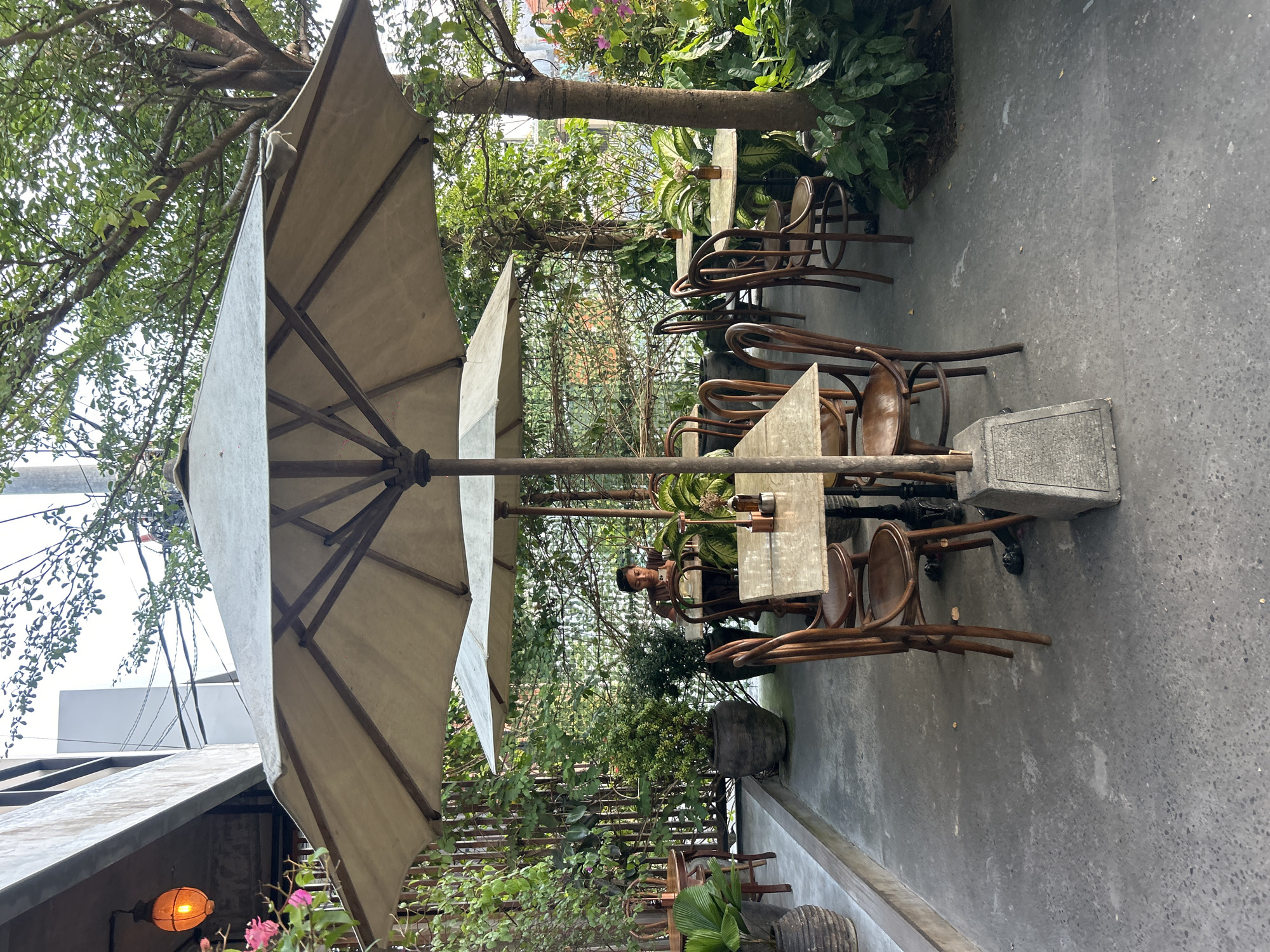 This image depicts an outdoor seating area of a cafe in North Kuta, Indonesia. Two large umbrellas provide shade over tables and chairs. A person is seated at a table in the background, looking at a phone. The area is surrounded by lush greenery, including trees and potted plants. The ground is paved with concrete. A lamp hangs from an overhang to the left, and a wooden fence is visible behind the plants.