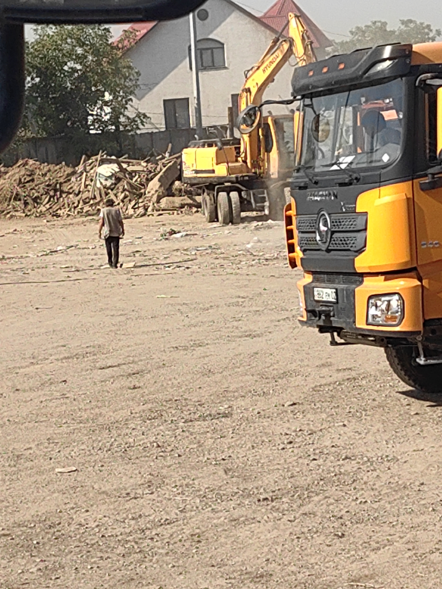 A person walks away from the camera on a dusty, unpaved lot. The person is wearing a light-colored vest over dark clothing. In the foreground, to the right, is the front of a yellow semi-truck. Behind the person is a large pile of debris, including wood and concrete. Further in the background, a yellow excavator is visible. To the left of the excavator is a light-colored building with a red roof. The sky is hazy. The scene appears to be a construction or demolition site.