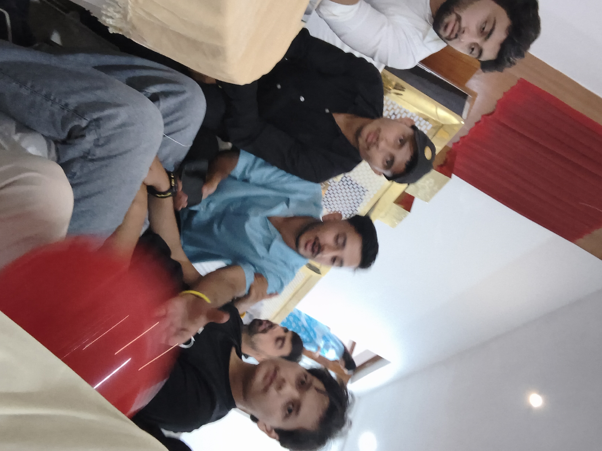 In the city of Khaniara, India, a group of five young men are gathered indoors, seated on the floor. The man on the far left wears a white shirt and has a beard. To his right, a man in a black shirt and a black baseball cap is visible. In the center, a man in a light blue shirt is speaking, with a red balloon obscuring parts of his body and the man to his right. The man to the right of the speaker wears a black t-shirt and has a yellow bracelet on his left wrist. The fifth man, partially obscured, is seated behind the man in the black t-shirt. The background features a red draped cloth and decorative golden paneling. The overall lighting suggests an indoor environment.