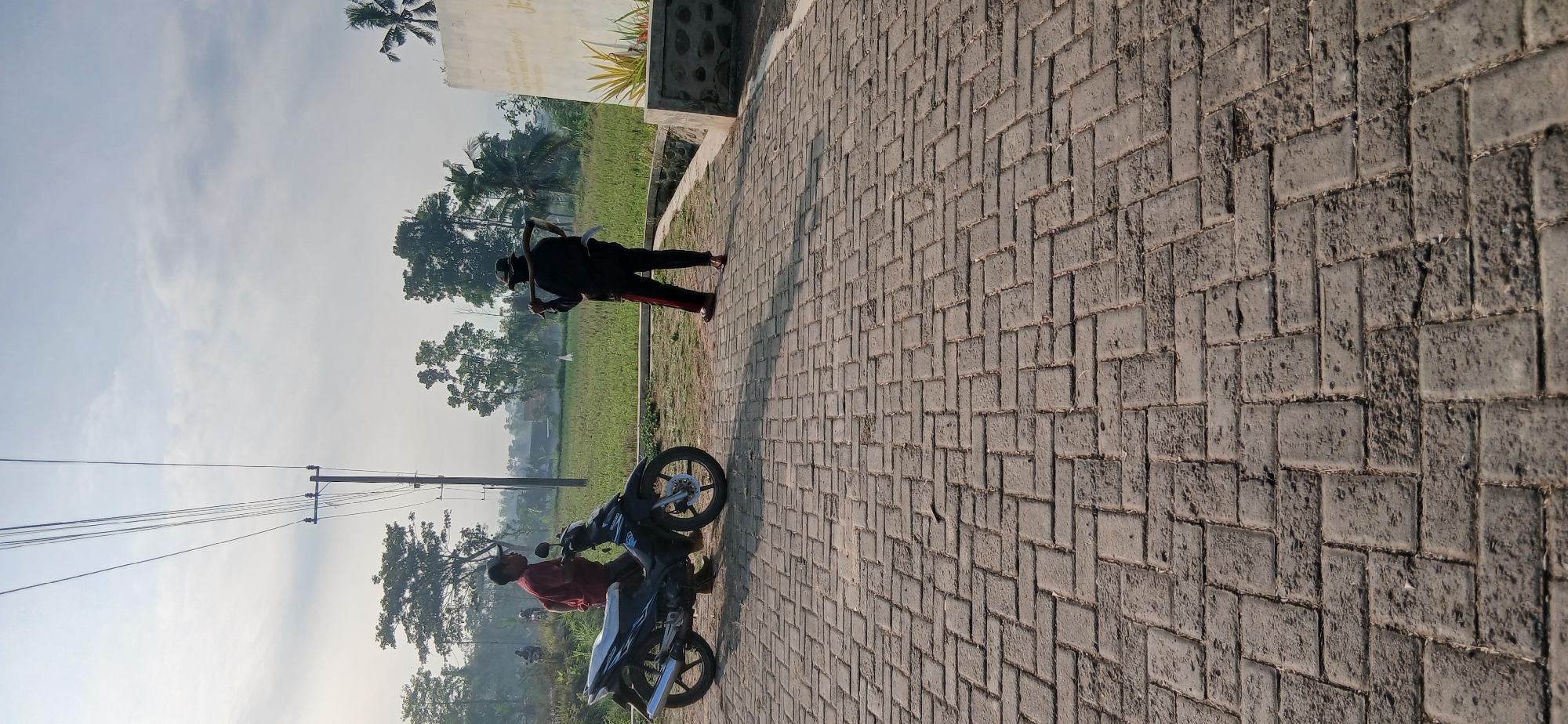 In Locare, Indonesia, on a sunlit day, two individuals are present on a paved walkway. A man wearing a maroon shirt and a dark baseball cap stands next to a parked black motorcycle. Across from him, another person dressed in black pants with red stripes, a black shirt, and a hat, carries a curved tool over their shoulder. Behind them, a field of green crops extends towards a treeline, with a telephone pole and wires visible in the distance. A white wall with some writing and a palm tree are situated on the right side of the frame. The ground is covered in interlocking gray pavers.
