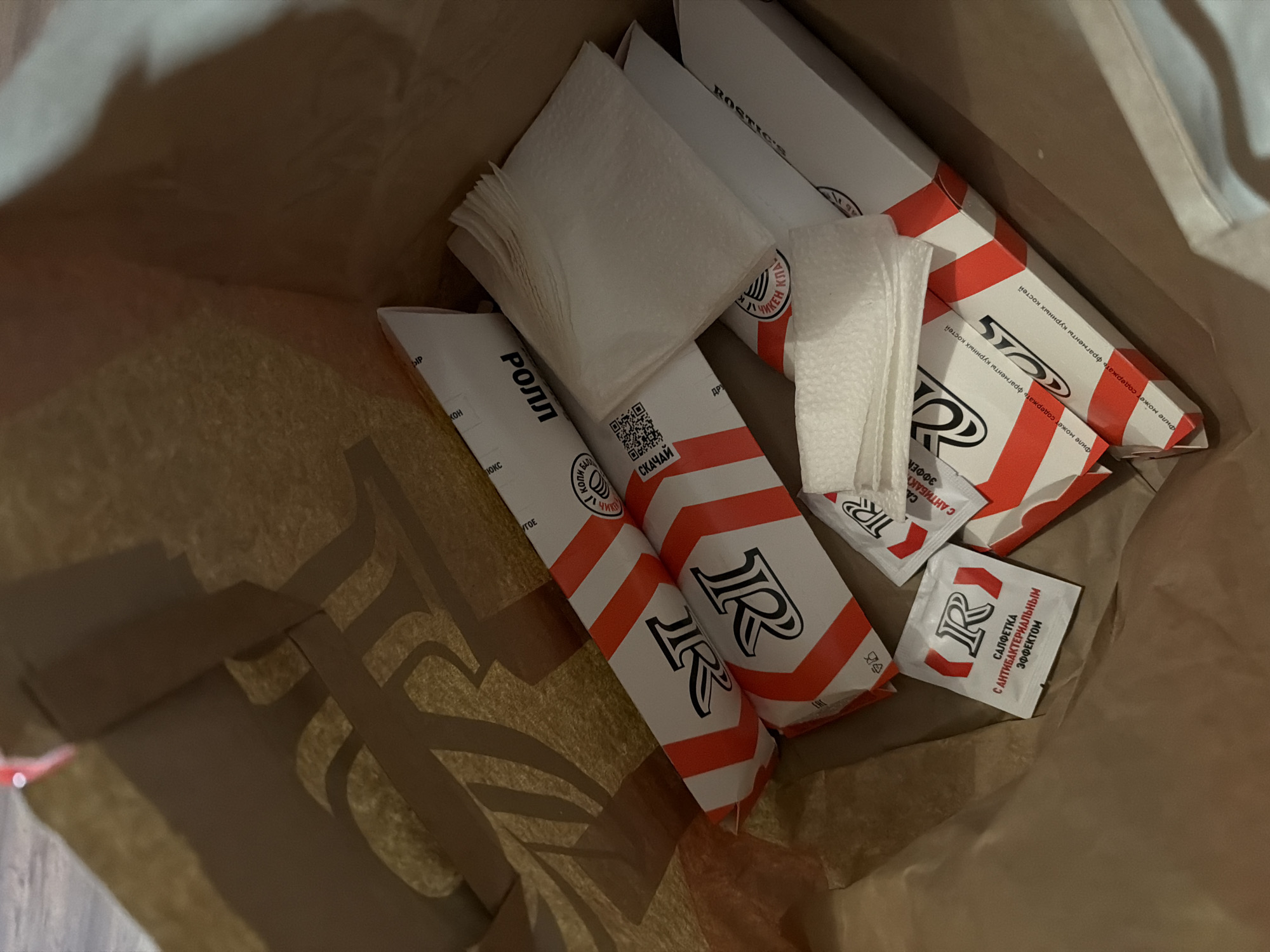 A view from inside a brown paper bag containing food items from the restaurant "Rostic's" (formerly known as KFC), possibly in Perm, Russia. Visible are packages of "ROLL" sandwiches, rectangular boxes of "ROSTIC'S" items, a stack of napkins, and individual packets of antibacterial wipes. The branding on the packaging and wipes features the "R" logo of Rostic's. The items are arranged within the bag.