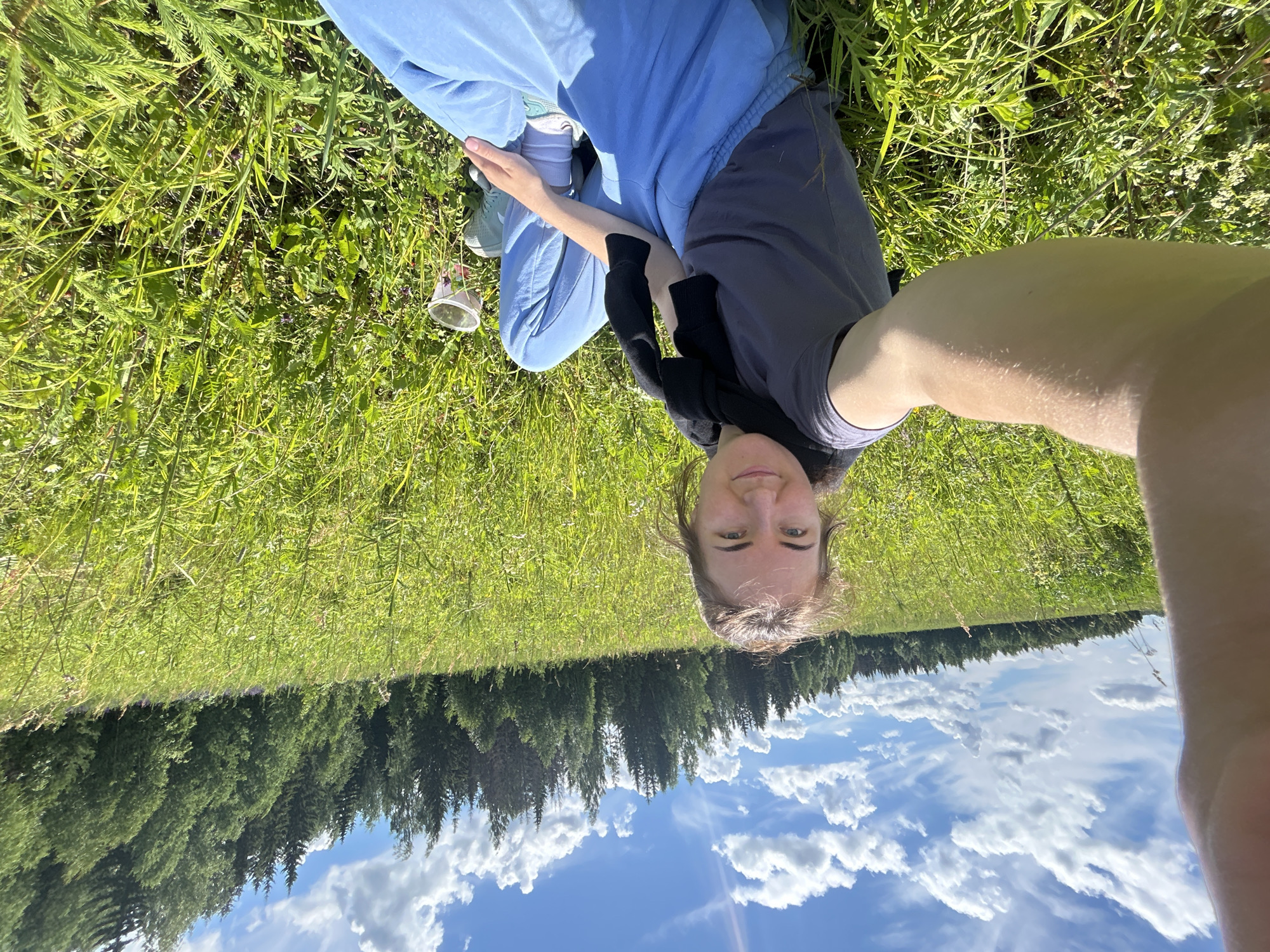 In the city of Shabury, Russia, a young woman is captured in a selfie taken outdoors in a grassy field. She is wearing a grey t-shirt, light blue sweatpants, and light blue sneakers. A black sweater is draped around her shoulders. She is sitting on the ground, with tall green grass surrounding her. In the background, a dense forest with tall evergreen trees forms a backdrop against a bright blue sky dotted with white, fluffy clouds. The overall scene suggests a pleasant, sunny day in a rural or natural setting.