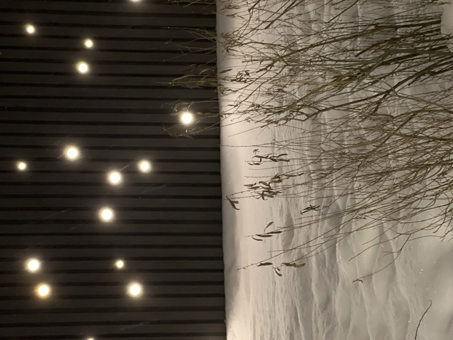 A stark contrast divides the image: a black background with evenly spaced, glowing orbs on the left and a snow-covered landscape with bare branches on the right. The branch tips extend slightly onto the black section. The lighting suggests twilight or nighttime conditions, and the snow appears untouched. The black background has horizontal lines, creating a patterned effect.