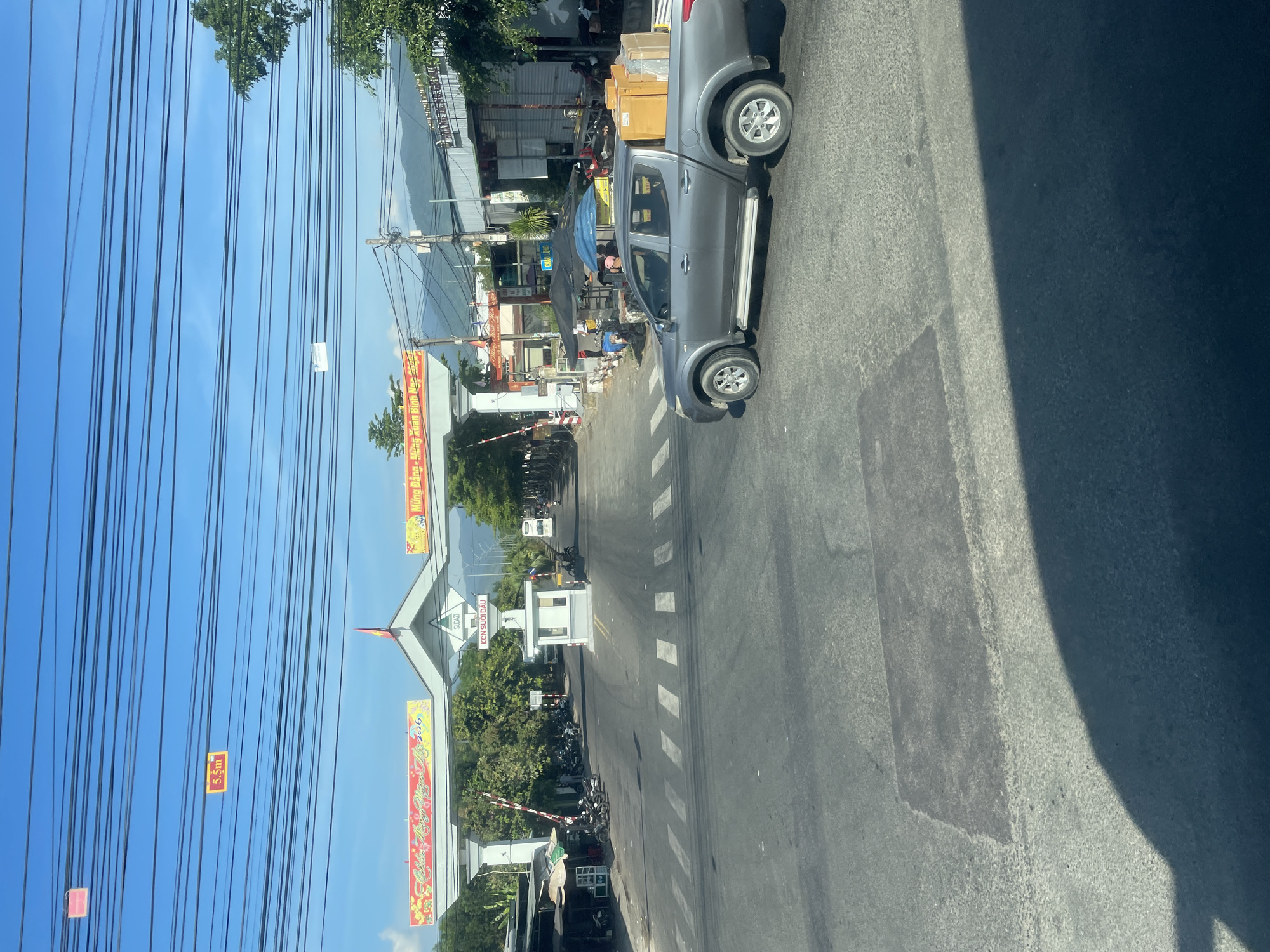 The image depicts a view from a vehicle looking towards a gateway with overhead power lines. The gateway features two arched structures, one with a banner displaying text in Vietnamese that appears to be a greeting and the year 2016. Beneath the arch, there is a booth with a barrier, and a road with pedestrian crossings. Various motorcycles are parked on the side of the road, and a grey pickup truck carrying boxes is moving through the scene. Buildings and trees are visible in the background, and mountains can be seen in the distance under a clear blue sky. The location is identified as Tay Nha Trang Ward, Vietnam.