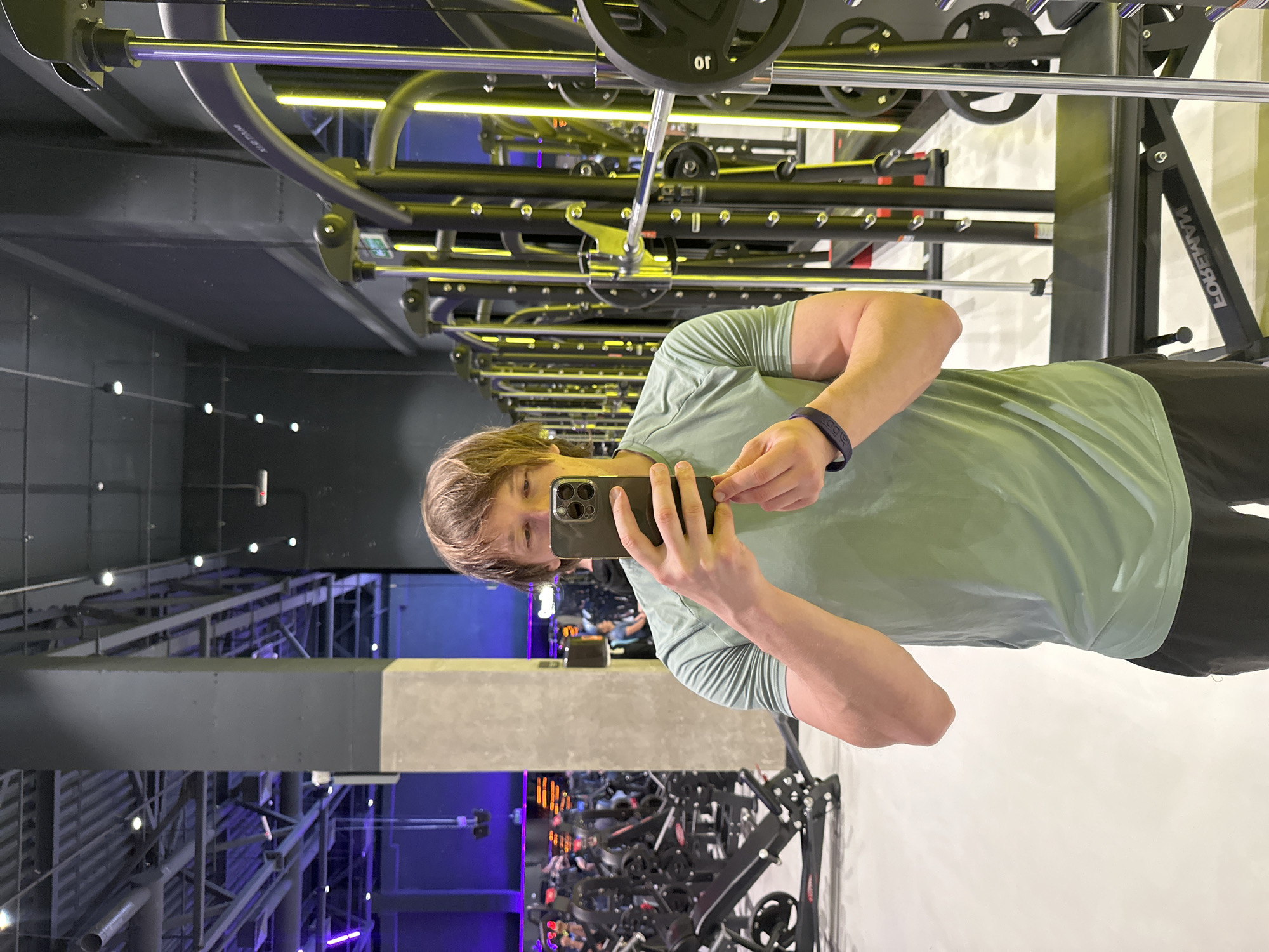 A young adult male is taking a mirror selfie in a brightly lit, modern gym. He is wearing a pale green t-shirt and dark shorts, with a dark blue wristband on his left wrist. The gym equipment, including weight machines and benches, is visible in the background, with yellow LED lights accentuating the equipment. The overall environment suggests a commercial fitness facility.