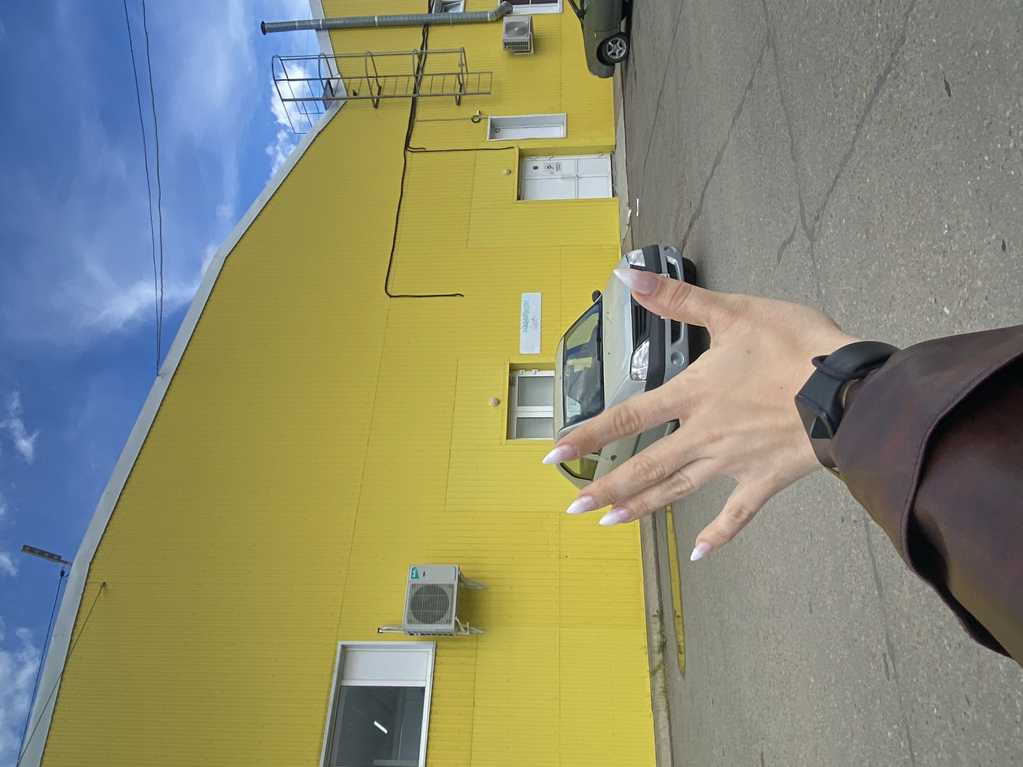 The media file captures a first-person perspective, primarily featuring a person's outstretched right hand with a bright yellow building and sky in the background. The image is taken outdoors on a clear day with a blue sky dotted with white clouds.
The subject's hand is in the foreground, extended slightly towards the center of the frame. It has long, almond-shaped nails with a white tip (French manicure style). A black smartwatch with a black strap is visible on the wrist, partially covered by a brown jacket or sleeve.
The main object in the background is a large, brightly colored yellow building, possibly an industrial or commercial structure like a warehouse or factory. Its exterior is made of corrugated metal panels. The building features several windows, white doors, and external fixtures such as a metal ladder leading up the side, a chimney or vent pipe on the roof, and two external air conditioning units mounted on the wall. Black electrical conduits or pipes run along the building's facade.
In front of the building, a white commercial van is parked, with its front visible. Part of a green car is also visible in the upper right corner, suggesting a parking area. The ground consists of asphalt pavement, showing cracks in some areas. A small, faded rectangular sign with illegible text is visible on the yellow wall.
The setting appears to be an urban or industrial area in Chaikovsky, Russia, characterized by functional architecture and paved surfaces. The bright lighting suggests it is daytime.