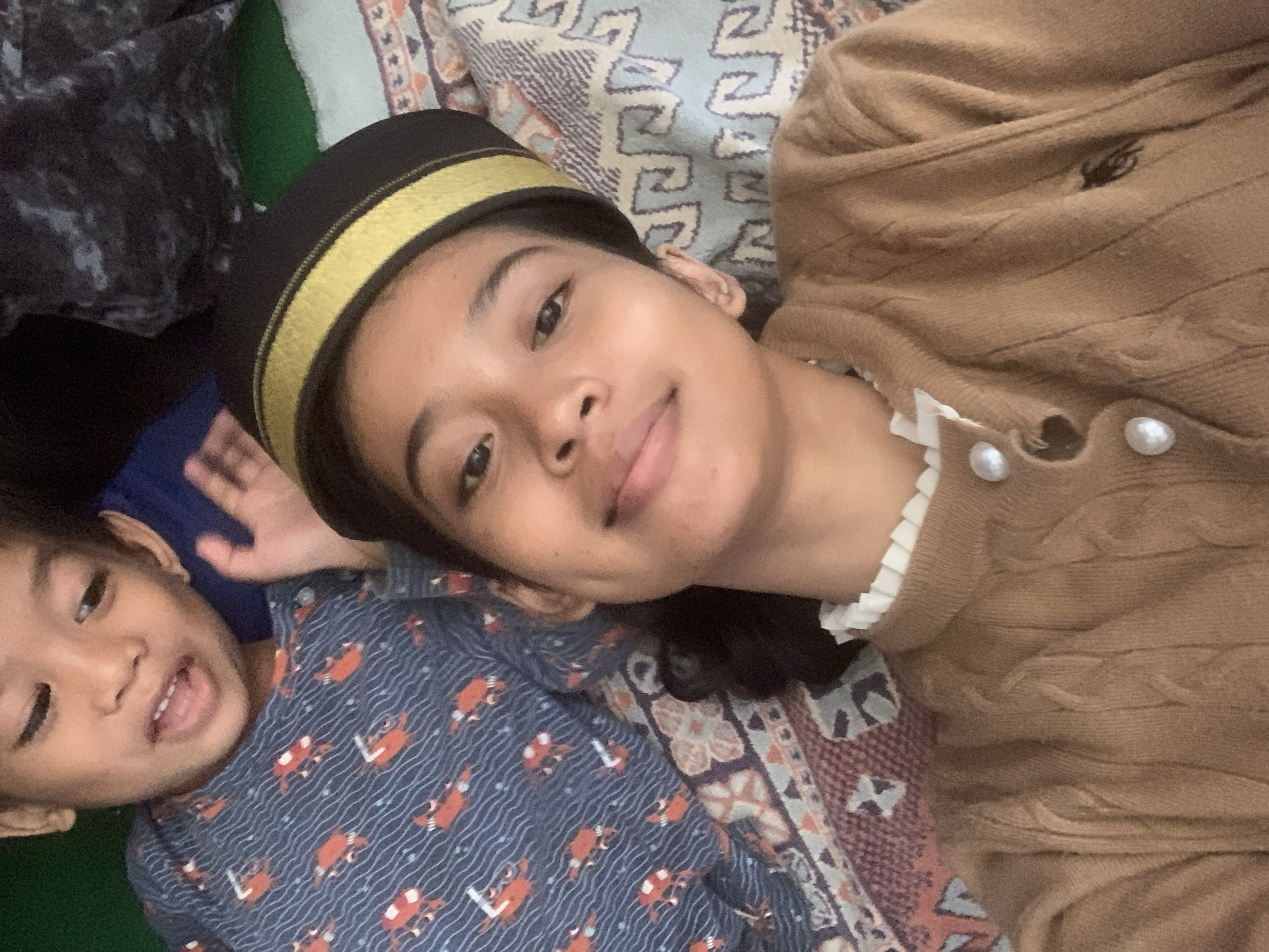 Two individuals, a child and a young woman, are lying down in what appears to be a bedroom in Sungai Meriam, Indonesia. The child, positioned in the upper left of the frame, is wearing a blue pajama top with an orange crab print. The young woman, in the foreground, is wearing a brown cardigan with a white ruffled collar and a black hat with gold trim. The background shows bedding with a patterned design. The scene is a close-up selfie, suggesting the photo was taken by the young woman.