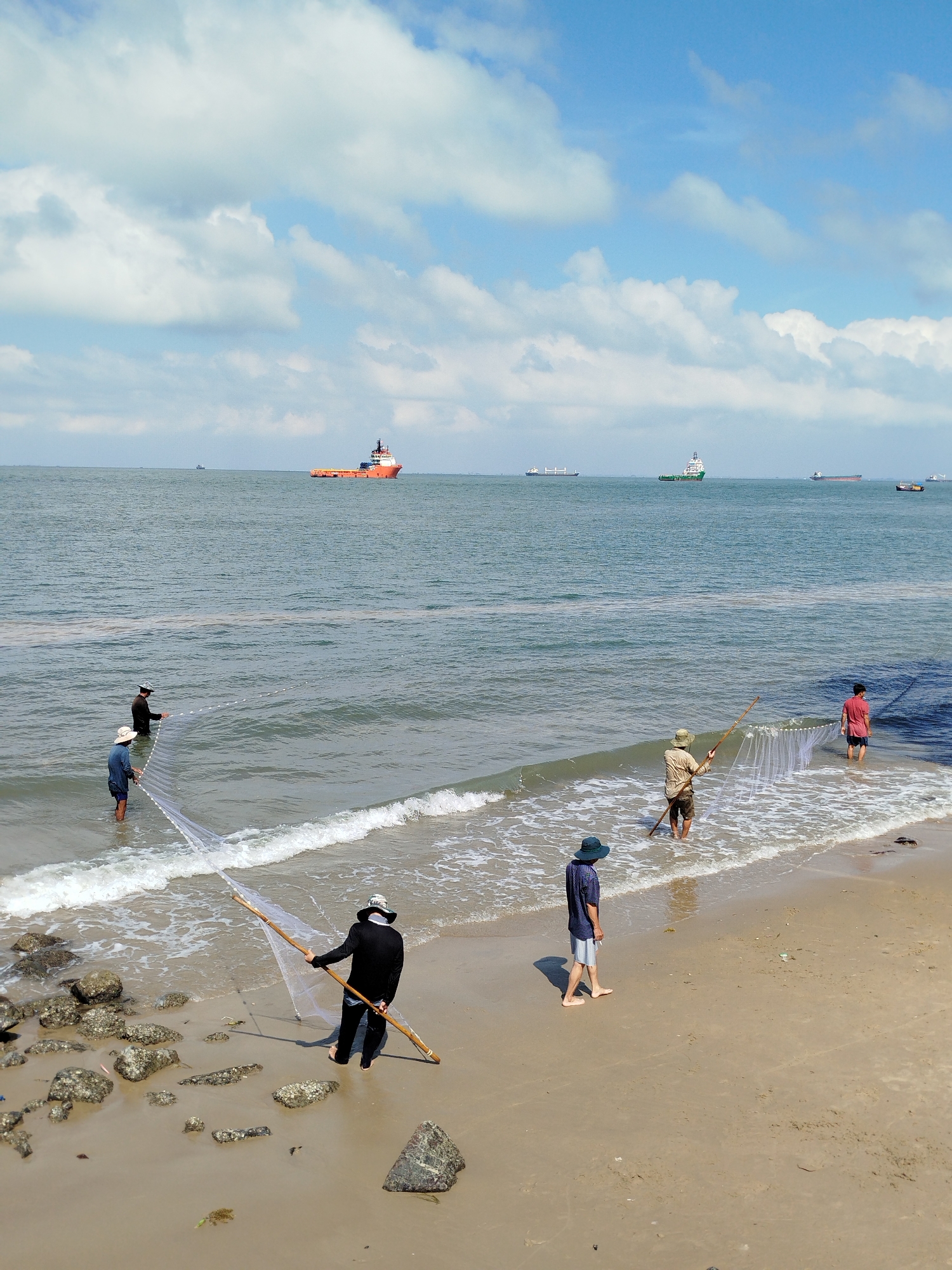 The photo depicts a group of people fishing on a beach in Ho Chi Minh City, Vietnam. Five individuals are present. Two men are standing in the water, holding a long net between them. Another man on the beach holds a long pole connected to a net. A fourth person is standing on the sand, and another is wading in the water. They are all actively involved in fishing.
The scene is set on a beach with a mix of sand and some rocks near the water's edge. The ocean is visible in the background with several ships on the horizon under a blue sky dotted with white clouds.