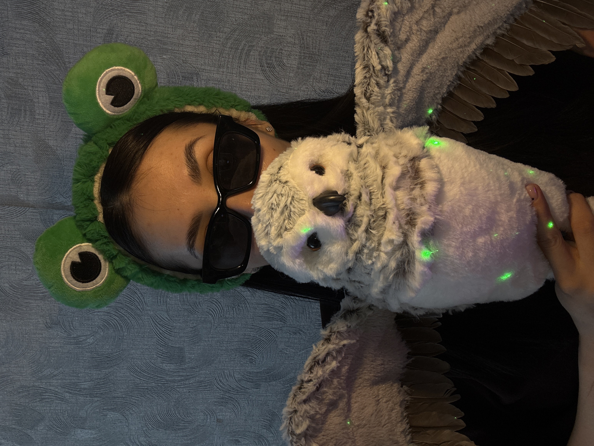 A person wearing sunglasses and a green frog-shaped hat lies on a blue patterned surface, holding a stuffed owl with wings. The location is not discernible from the image and Votkinsk, Russia, cannot be determined.