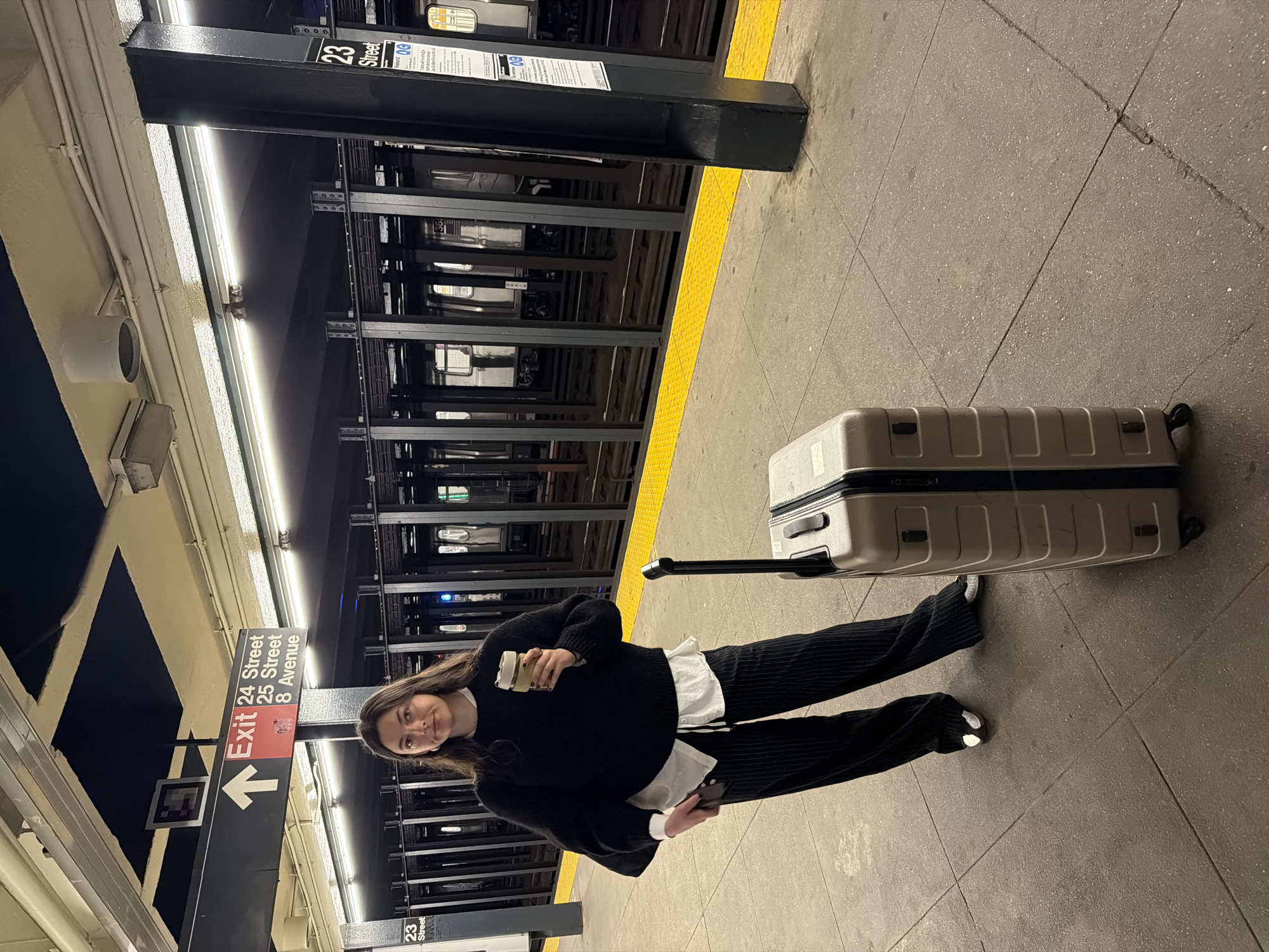 The image depicts a woman standing on a subway platform in New York City, United States of America. She is holding a coffee cup, a phone and a suitcase. The signs nearby indicate the 23rd street station in midtown Manhattan. An "Exit 24 Street/25 Street/8 Avenue" sign points towards an exit. The station has yellow safety markings along the platform edge. A train is visible behind the safety barrier. The lighting in the station is provided by fluorescent lamps.