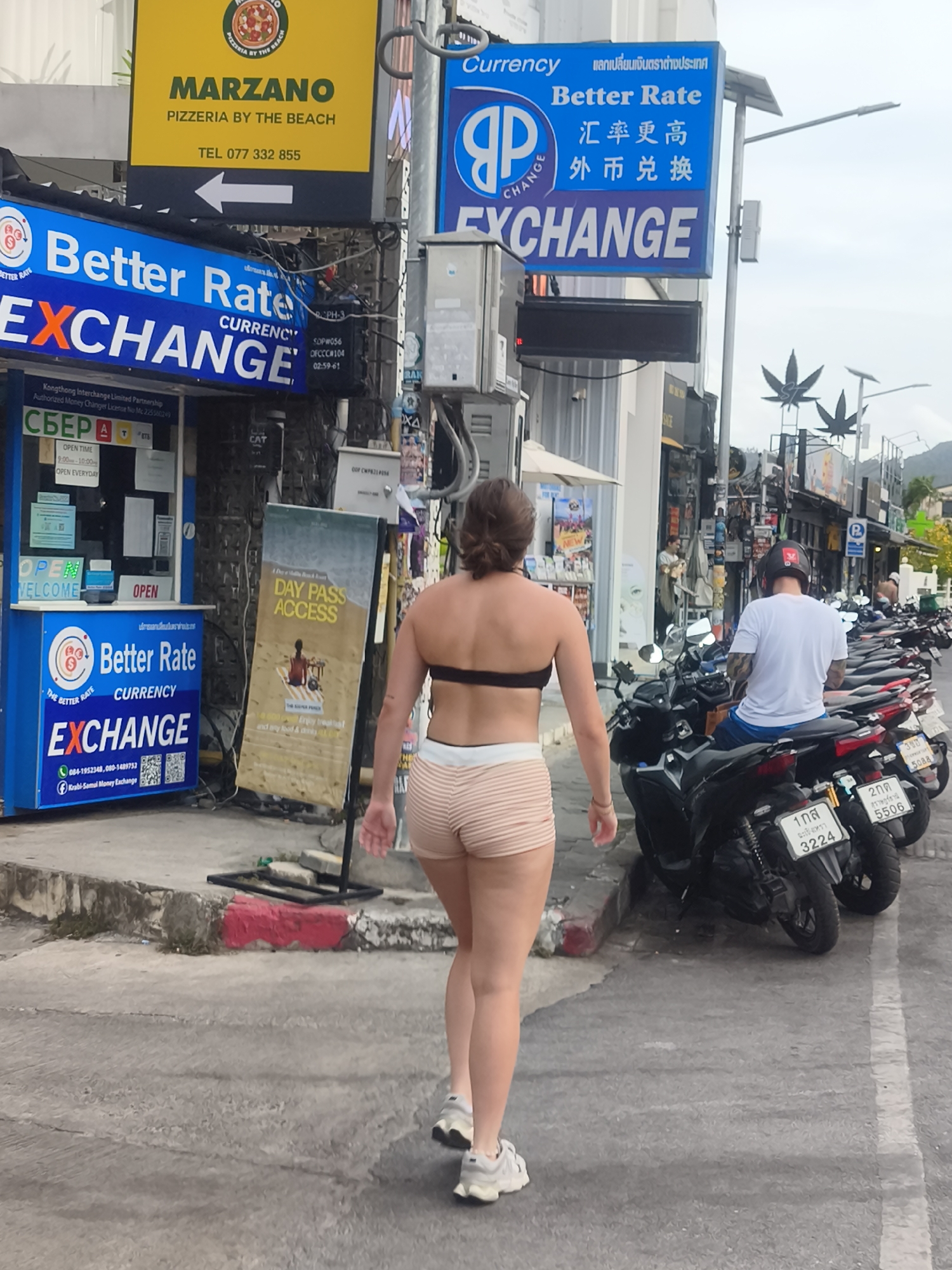 In a street scene in Ko Samui District, Thailand, a woman walks away from the camera. She is wearing a black bikini top and striped shorts, with athletic shoes. To her left is a blue storefront displaying "Better Rate Currency Exchange" and "OPEN WELCOME." Behind her is a row of parked motorcycles. A yellow sign for "MARZANO PIZZERIA BY THE BEACH" is visible further up the street. The environment appears to be a commercial area with sidewalks and street infrastructure.