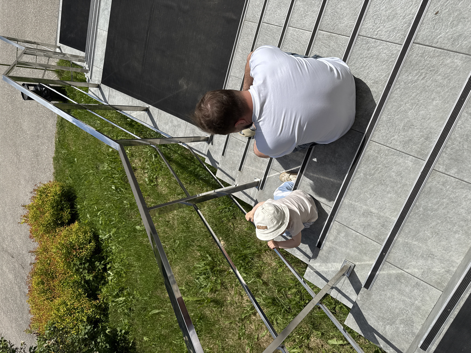 An overhead shot captures an adult and a young child on a modern outdoor staircase in Dobrograd, Russia, on a bright, sunny day. The stairs are made of grey tiles, each tread featuring a dark, ridged anti-slip strip. A sleek, shiny metal railing runs alongside the stairs.
The adult, a person with short brown hair, is seen from behind, wearing a white t-shirt and light-colored shorts or pants, kneeling or crouching on the stairs. They appear to be looking down towards the child. Adjacent to the adult, further down the stairs, a small child is also crouching, holding onto the metal railing with both hands. The child wears a light-colored t-shirt and a light-colored bucket hat. The word "BROWN" is visible in an arc across the front of the child's hat. Both subjects cast distinct shadows, indicating strong, direct sunlight, suggesting it's likely midday.
To the left of the stairs, there is vibrant green grass and several green and reddish-brown bushes. Beyond the foliage, a paved area, possibly an asphalt path or road, is visible. The scene conveys an interaction between the adult and child, possibly an act of supervision, teaching, or shared exploration on the stairs.