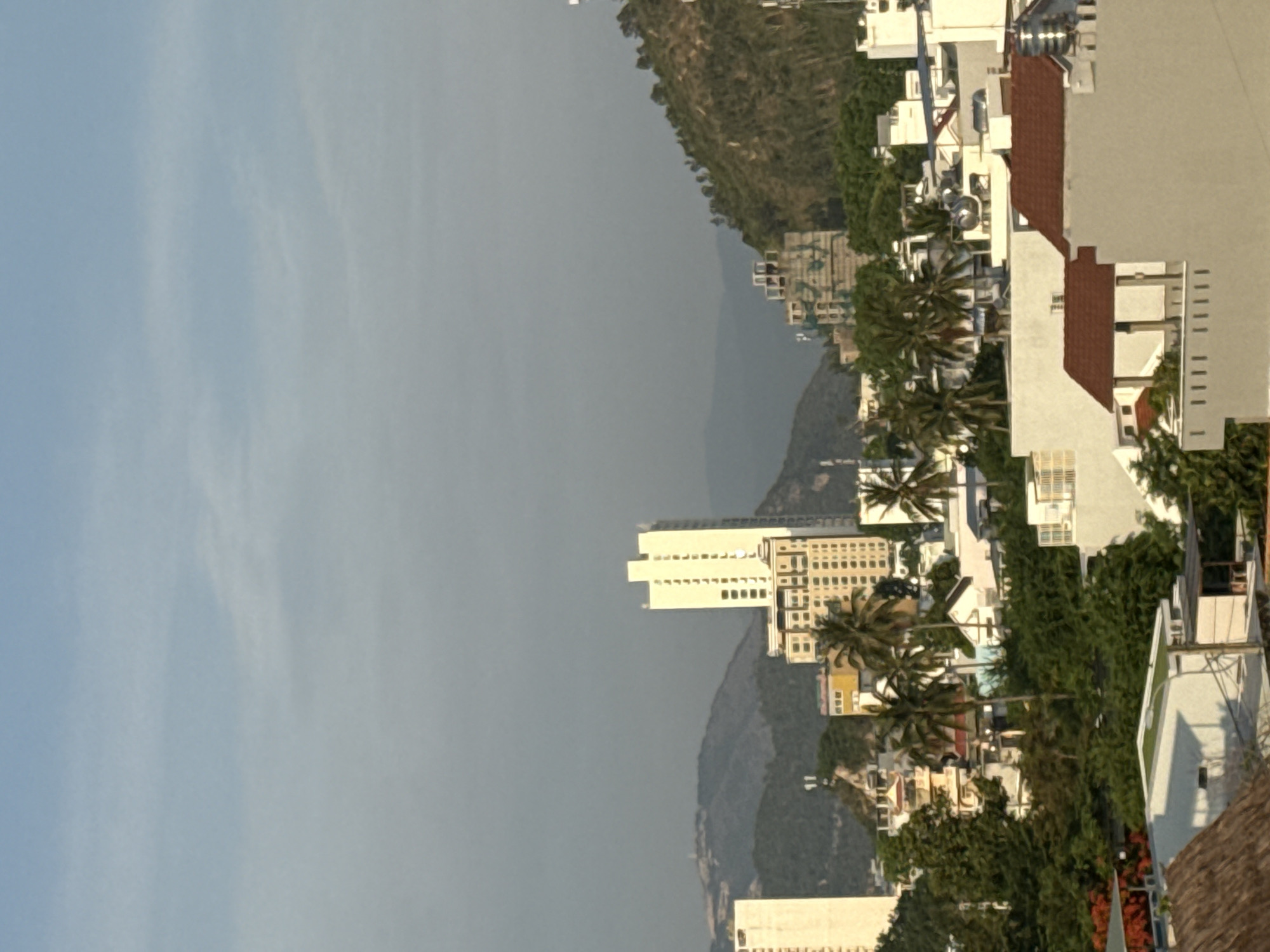 The image shows a cityscape with buildings and lush greenery under a hazy sky. In the foreground, several white buildings with red-tiled roofs are visible, some appearing residential with outdoor living spaces. Further into the background, taller, modern buildings stand out, including a prominent white high-rise with many windows and balconies. There are also a few smaller, older buildings with more intricate architectural details. Palm trees are scattered throughout the scene, adding to the tropical feel. In the distance, rolling hills and mountains create a natural backdrop to the urban development. The overall impression is that of a city nestled amongst nature, possibly experiencing a warm day with some atmospheric haze.