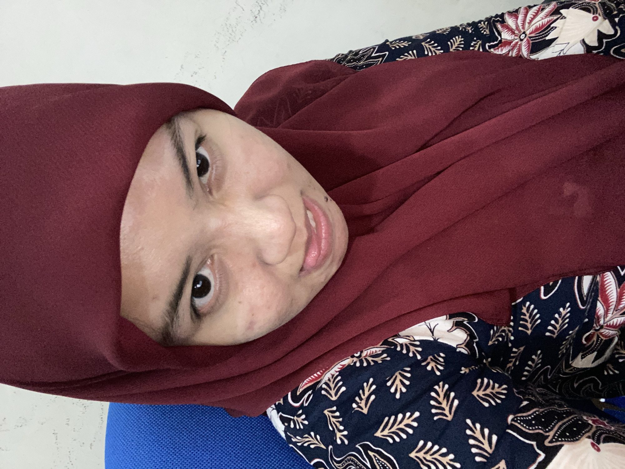 A young woman wearing a maroon hijab and a dark blue batik-patterned shirt is captured in a close-up, high-angle selfie. She is seated on a blue chair against a white wall. Her face is turned towards the camera, with her tongue slightly protruding and her eyes looking upward. The lighting is bright, originating from the front. The image quality is clear, with no visible artifacts. The background is plain and uncluttered, suggesting an indoor setting such as an office or a room. The overall impression is that of a candid or informal portrait.