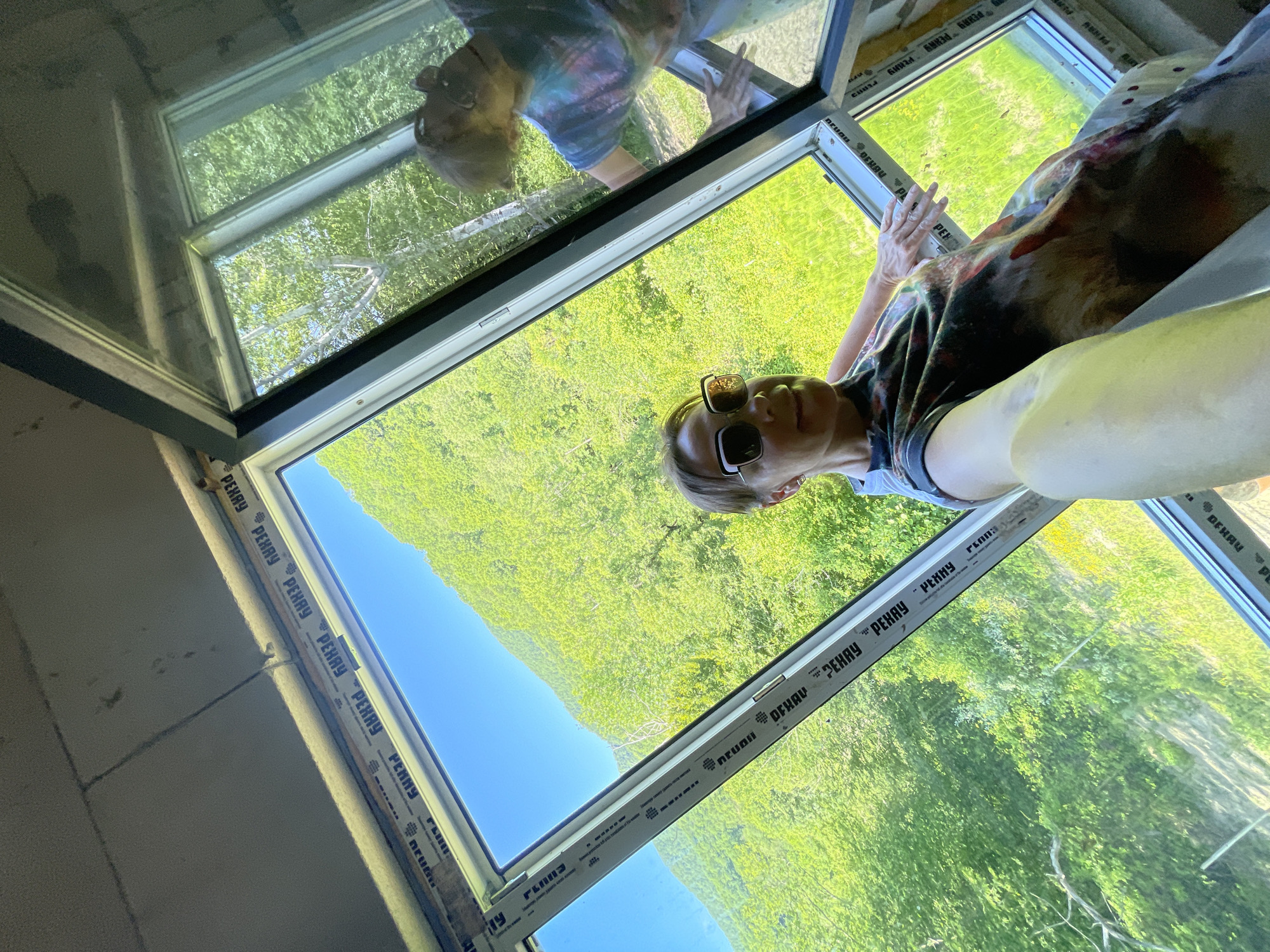 In Verkhniy Yurt, Russia, a woman is taking a selfie in a room with large windows offering a view of a lush, green hillside under a clear blue sky. She is wearing sunglasses and a colorful, abstract-patterned t-shirt. Her left hand is reaching out towards the window frame. The window frames have visible "PERRY" branding. Another person, partially visible in reflection on the window, is wearing a tie-dye shirt and appears to be cleaning the glass. The interior of the room is unfinished, with visible ceiling insulation and construction materials.