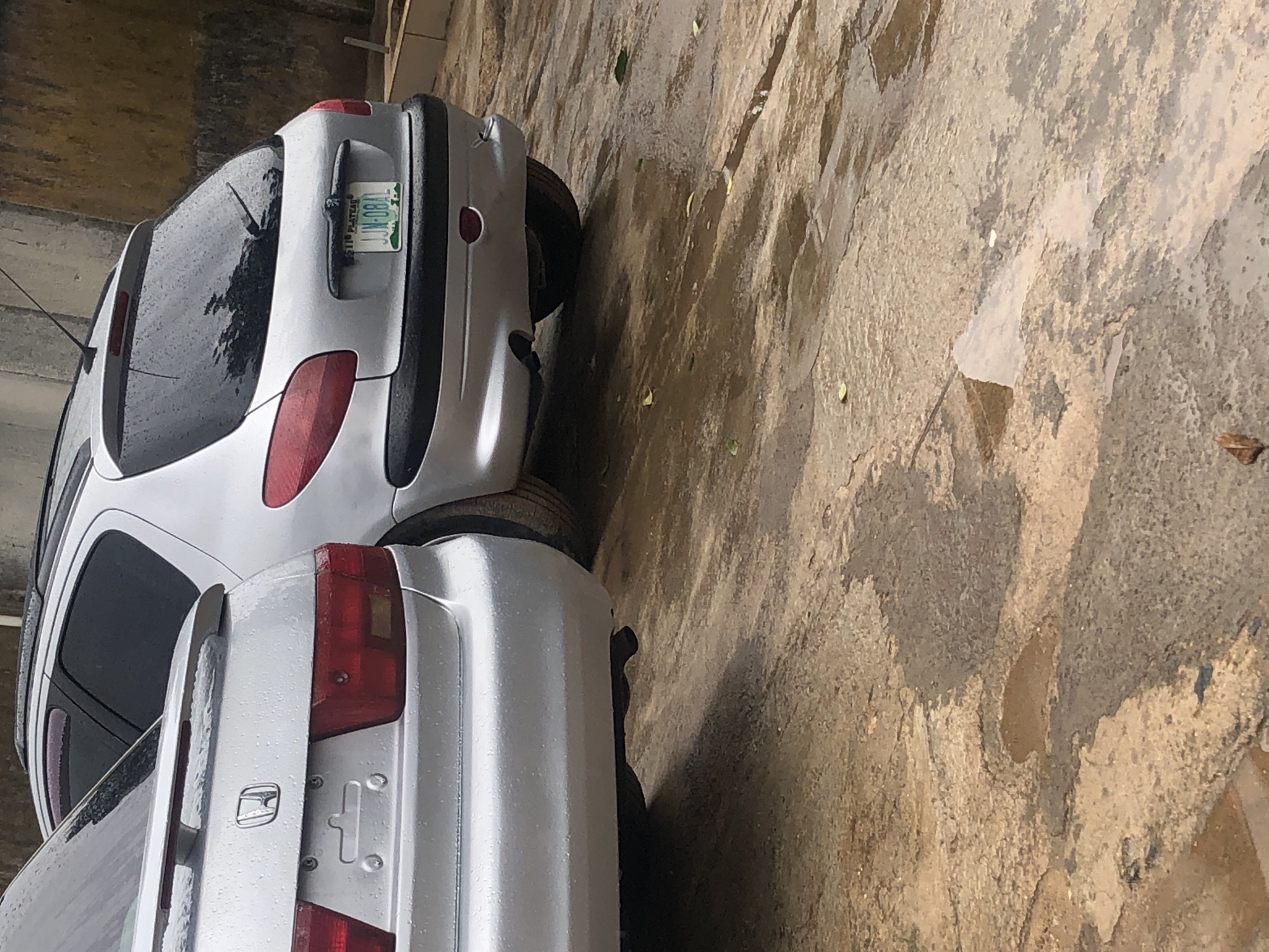 Two cars are parked on a wet, concrete surface. The car in the foreground is a silver sedan, partially visible from the rear quarter panel. The car in the background is a silver hatchback with a license plate that reads "JJN-0841". The ground is covered in puddles and scattered leaves, indicating it has been raining recently. The wall in the background is textured and appears to be made of concrete. The overall scene suggests an outdoor parking area in a city.