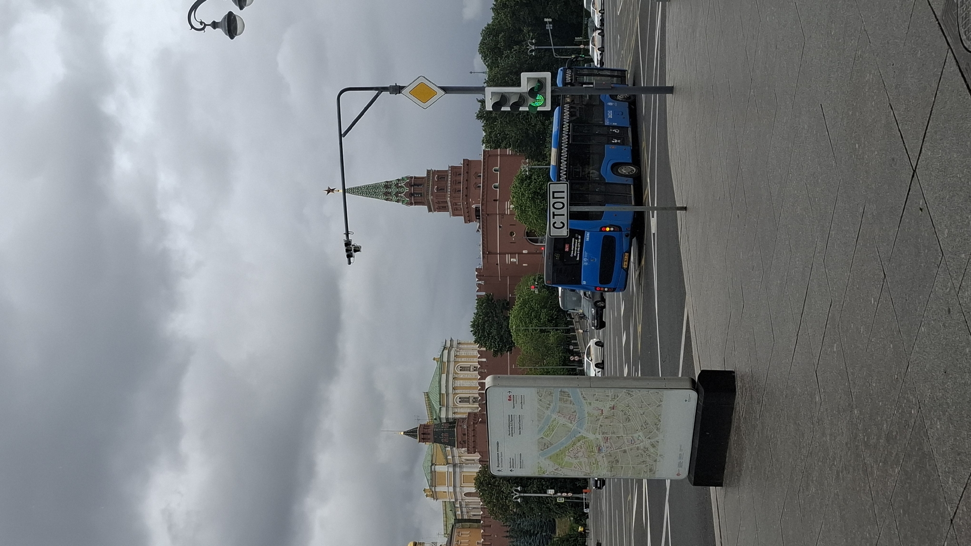 This is a vertically oriented image showing a street scene in Moscow, Russia, on an overcast day. The dominant structure in the background is a tall, red brick tower with a green, spired roof topped with a star, likely part of the Kremlin. In the mid-ground, there are more buildings with green roofs and ornate facades, also typical of Moscow architecture.
A blue bus is stopped at a traffic light, with a sign in Russian on its side reading "СТОП" (STOP). The traffic light is green. The bus has Russian license plates. In the foreground, there's a paved pedestrian area with a map displayed on a stand. The map appears to be a city map of Moscow. The overall mood is subdued due to the cloudy weather. The time of day is likely daytime, given the lighting conditions.