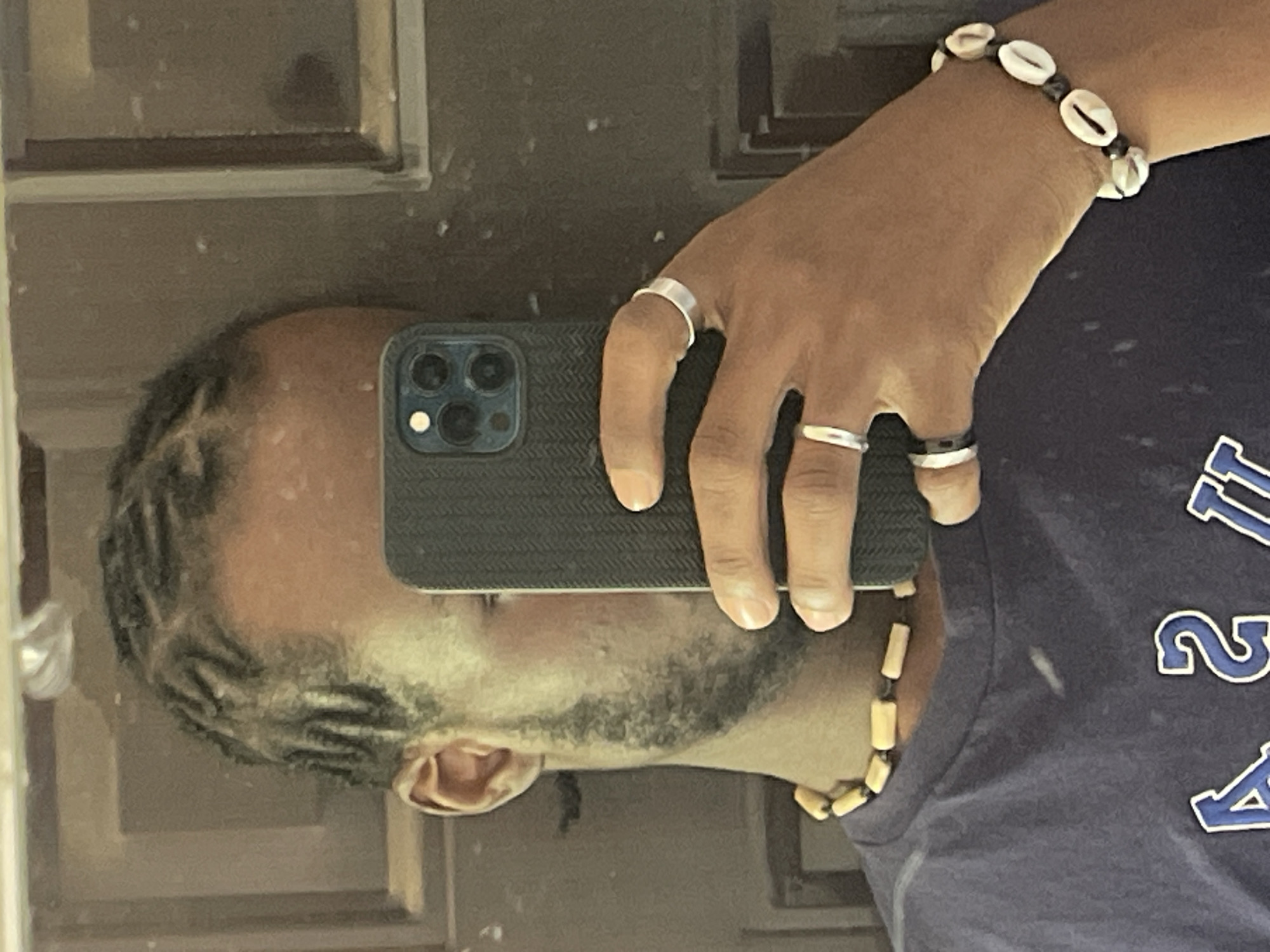 A young Black man, with braided hair, takes a mirror selfie in what appears to be an indoor environment, possibly a home, with a wooden door visible in the background. He is wearing a dark blue t-shirt with lettering that includes "USA" in blue and white. Around his neck is a beaded necklace of light-colored cylindrical beads. On his right hand, he wears two silver rings, one on his index finger and another on his ring finger. On his left wrist, he has a bracelet made of white cowrie shells and black beads. The man is holding a dark-colored smartphone in his right hand, obscuring the lower half of his face. The overall lighting suggests a moderately lit space. There are no specific indicators within the image that would allow for the identification of the city of Jos, Nigeria.