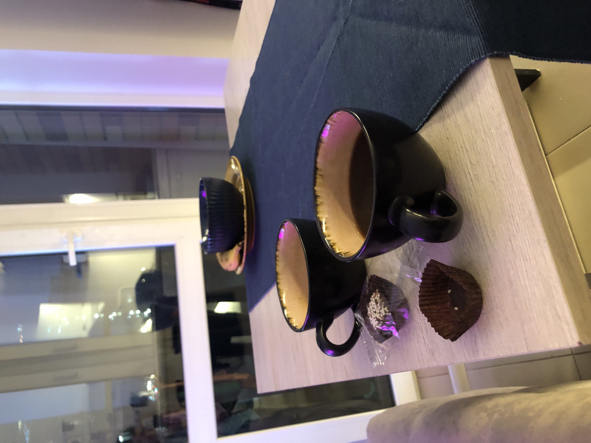 The scene depicts an indoor setting, specifically a light-colored wood-grain table with a dark blue fabric table runner, located in Saint Petersburg, Russia. On the table, two black ceramic mugs with textured tan/gold rims contain dark liquid, likely coffee. One mug is positioned closer to the viewer on the right, and the other is slightly to its left and further back. Between these mugs, two small dark chocolates in ruffled paper cups are present. One chocolate, wrapped in clear plastic, is topped with white sesame seeds; the other paper cup appears empty. In the background, on the table runner, a smaller dark blue ribbed bowl rests on a yellow-brown saucer. The background features a large white-framed window. The window glass reflects interior light sources and elements, including blurred white and purple lights, indicating an evening or nighttime setting. No individuals are visible in the image.