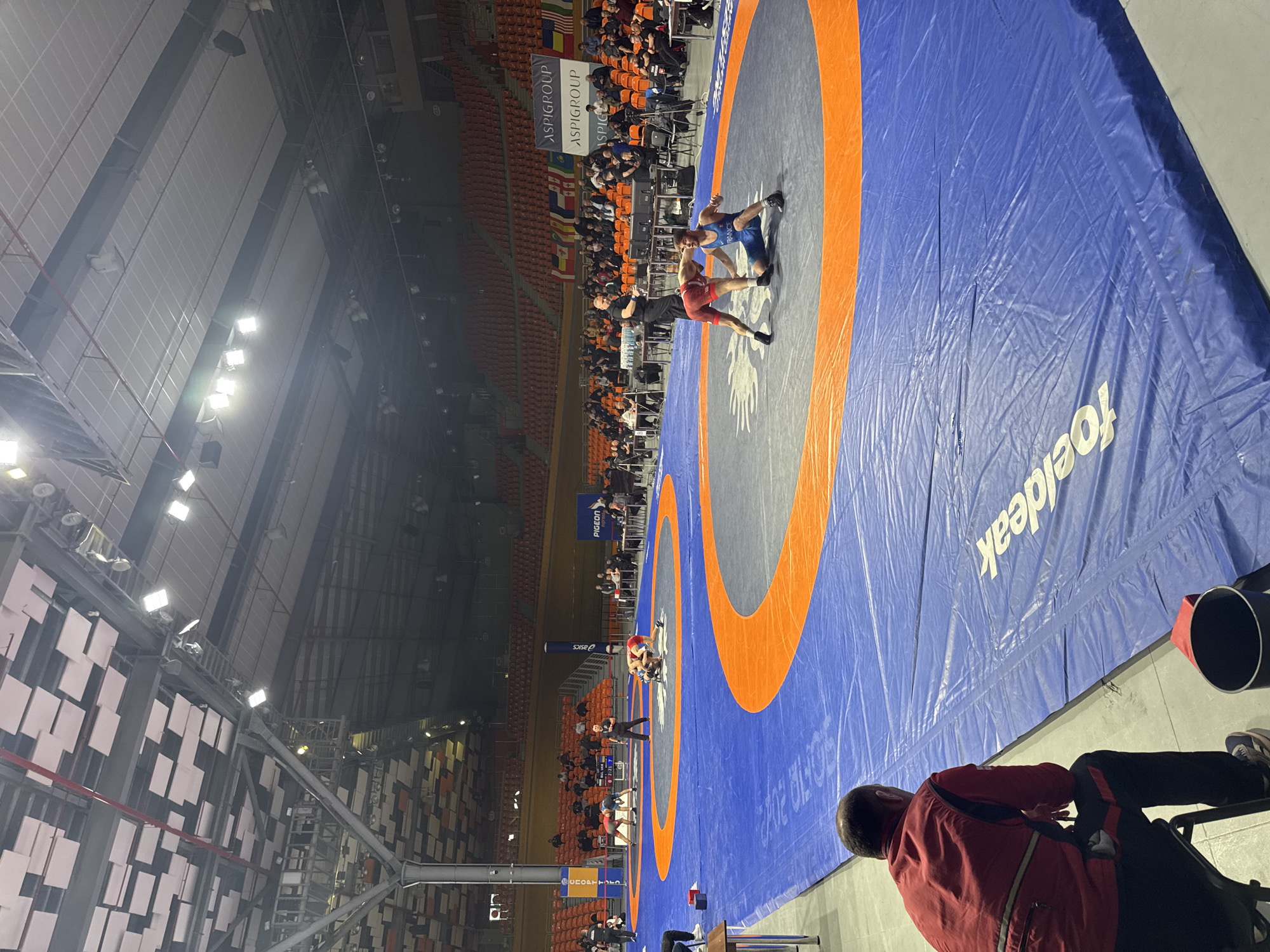 The scene depicts an indoor wrestling competition in progress at the Kolodrum Plovdiv arena in Plovdiv, Bulgaria. The spacious venue features a high ceiling with exposed structural elements and numerous overhead floodlights providing illumination. Tiered orange seating for spectators lines the perimeter, with banners displaying "ASPIGROUP", "PIGEON", and a series of national flags visible in the background.
At least three wrestling mats are laid out on a light grey concrete floor. The primary mat in the midground consists of a blue outer area, an orange inner ring, and a central grey circle adorned with a white sun-like emblem. On this mat, two male wrestlers are engaged in a match: one in a red singlet stands, leaning forward, while the opponent in a blue singlet kneels, looking up. A male referee in a black shirt and dark pants stands nearby, observing the wrestlers. A second mat to the left features two wrestlers in dark singlets grappling, supervised by another referee. A digital scoreboard to the left of the second mat partially displays "BUL UKR 0 2:21 7", indicating a match between Bulgaria and Ukraine with a score of 0-7 and a time of 2:21.
In the immediate foreground, a male, viewed from behind, is seated on a black chair. He wears a red long-sleeved jacket with black accents and dark pants, facing the wrestling mats. A black bucket with a red interior rim rests on the floor beside him. Numerous individuals, identified as officials or coaches, are seated at tables positioned along the edges of the mats, monitoring the ongoing matches. Spectators are sparsely distributed throughout the tiered seating.