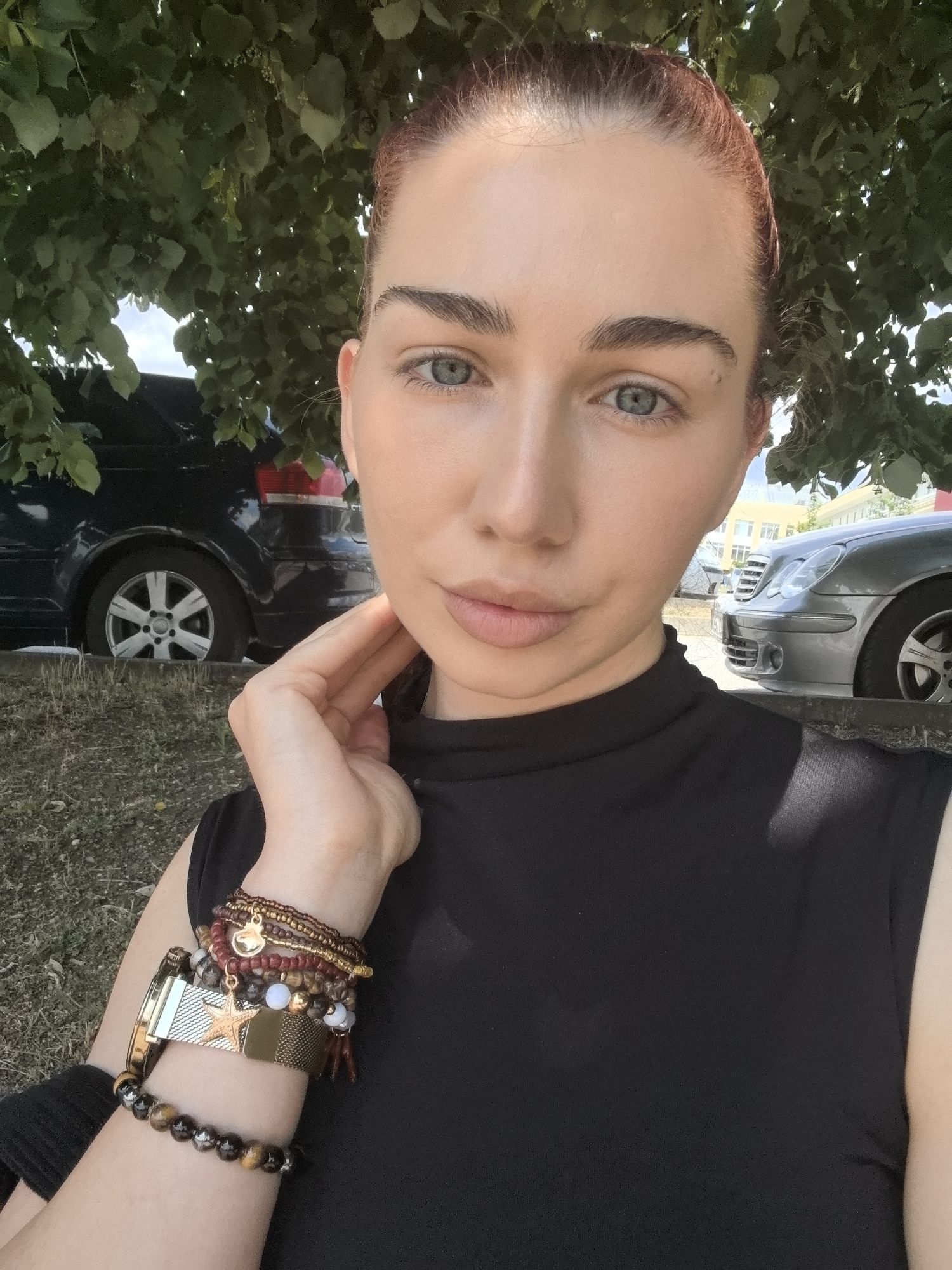 A daylight outdoor portrait features a woman with light skin, blue eyes, and reddish-brown hair pulled back, captured from the shoulders up. She looks directly at the camera with a neutral expression, her head slightly tilted. Her right hand is gently placed against her neck and jawline. She wears a black, high-necked, sleeveless top. Her left arm is adorned with several layered bracelets, including beaded strands in earthy tones, metallic bands, and charms, one of which is a prominent star shape and another a small heart or bell. A gold-toned watch is also visible on her left wrist.
The setting appears to be a street or parking area in Bogomilovo, Bulgaria, under the shade of a large tree with dense green foliage, casting dappled light. In the background, to the left, the rear of a dark blue car is partially visible, parked on a grassy or dirt verge next to a paved area. On the right, the front and side of a silver sedan are visible, also parked, with some distant buildings under a bright sky. The clear light suggests a sunny day. No text is visible in the image.