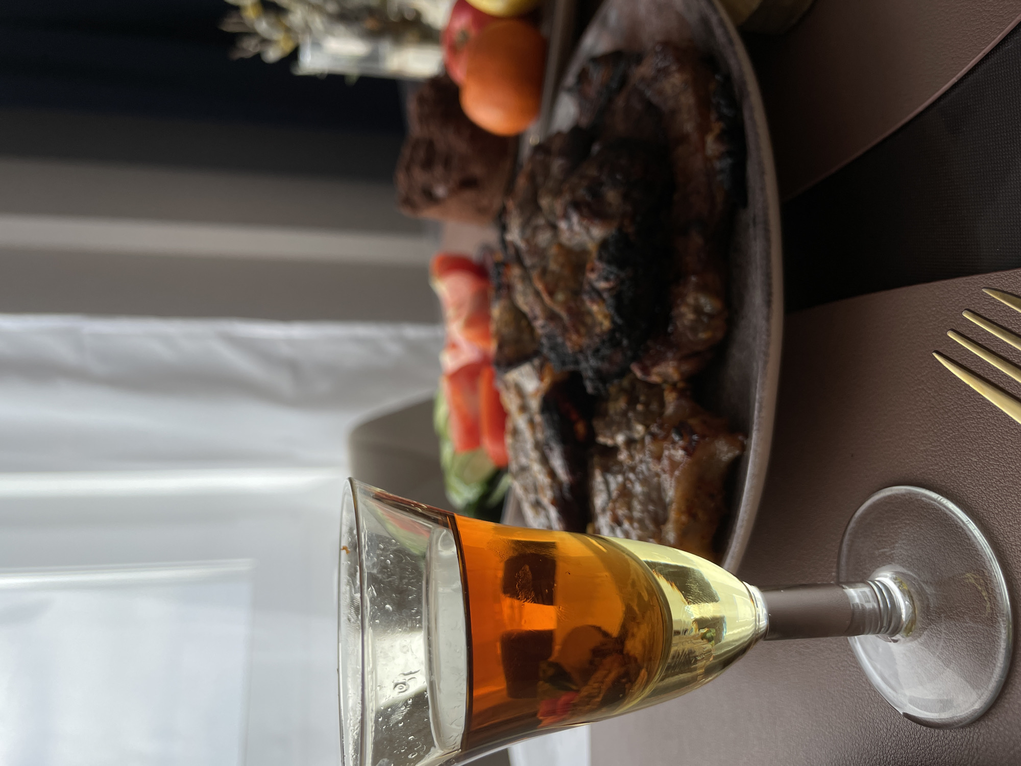 A still life is presented in a slightly canted, medium shot featuring a glass of amber liquid, likely wine or sherry, resting on its side in the foreground. Behind it, a wooden platter holds a pile of grilled meat, accompanied by fresh tomato and cucumber slices. Apples are partially visible at the back of the platter, adding to the arrangement. A set of golden-toned cutlery is partially seen on the right side of the frame, suggesting a dining setting. The backdrop consists of a window with light-colored drapes and a dark panel above, possibly indicating an indoor restaurant ambiance. The location in the city of Подшивалово, Russia, cannot be verified from the image content.