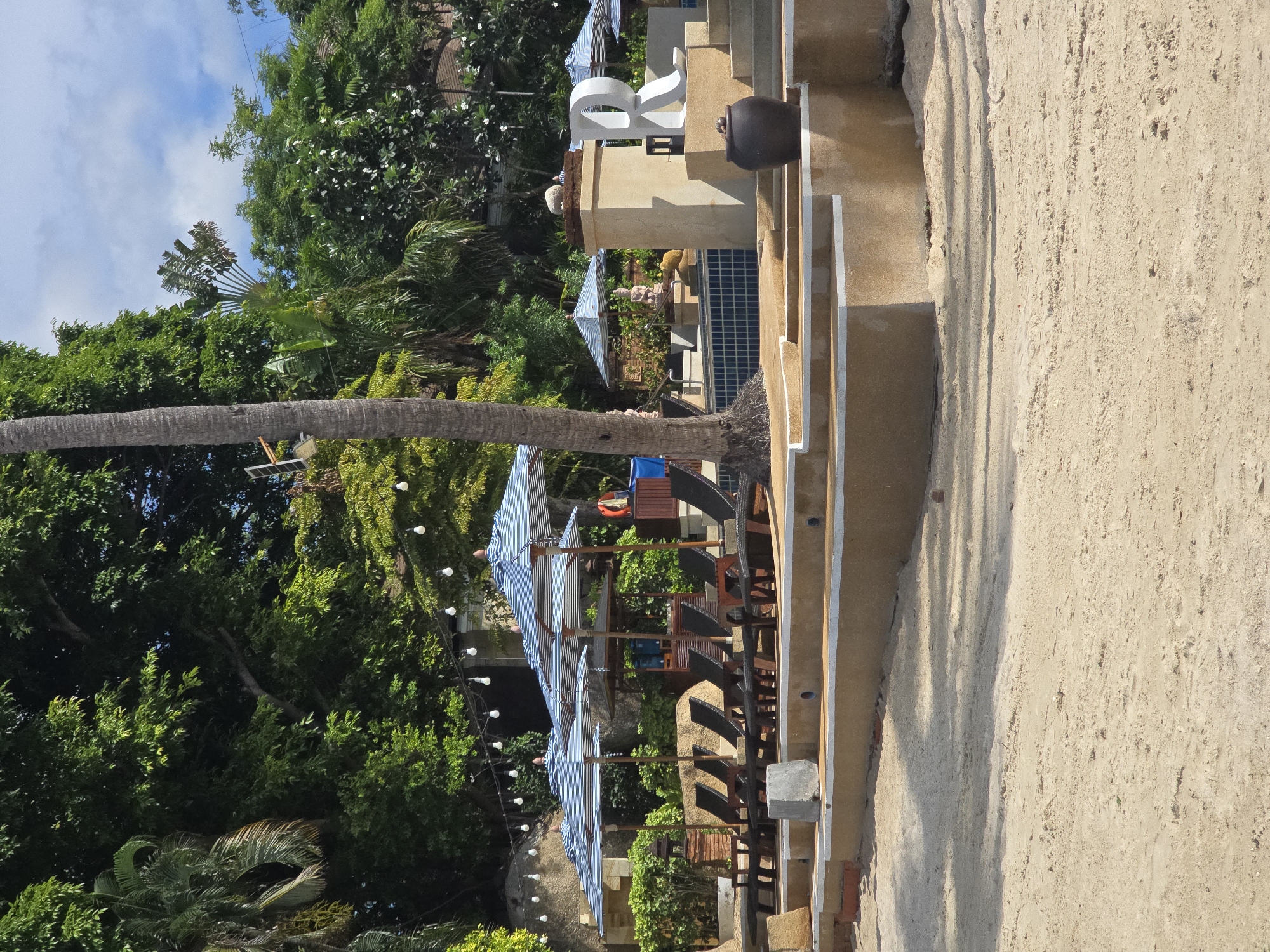 The image depicts a tropical beachside resort area in Baan Chaweng Noi, Thailand. In the foreground, light-colored sand stretches across the bottom of the frame. Behind the sand, a tiered concrete and stone structure rises, featuring stairs and ledges. On one of these ledges, a large, white, three-dimensional letter "R" stands prominently. To the left of the "R", a large, dark, ceramic pot is placed.
Further back, a swimming pool with blue tiled flooring is visible. The pool area is furnished with several rows of empty, dark-colored lounge chairs, some covered by blue and white striped umbrellas. Behind the lounge chairs, a variety of lush green tropical trees and foliage form a dense backdrop. Hanging string lights with exposed bulbs are suspended across the upper portion of the scene, suggesting an evening ambiance or decoration. A tall palm tree trunk is present on the left side of the frame, partially obscuring the view of the trees behind it. The sky above is partly cloudy. The overall scene conveys a serene and inviting resort atmosphere.
