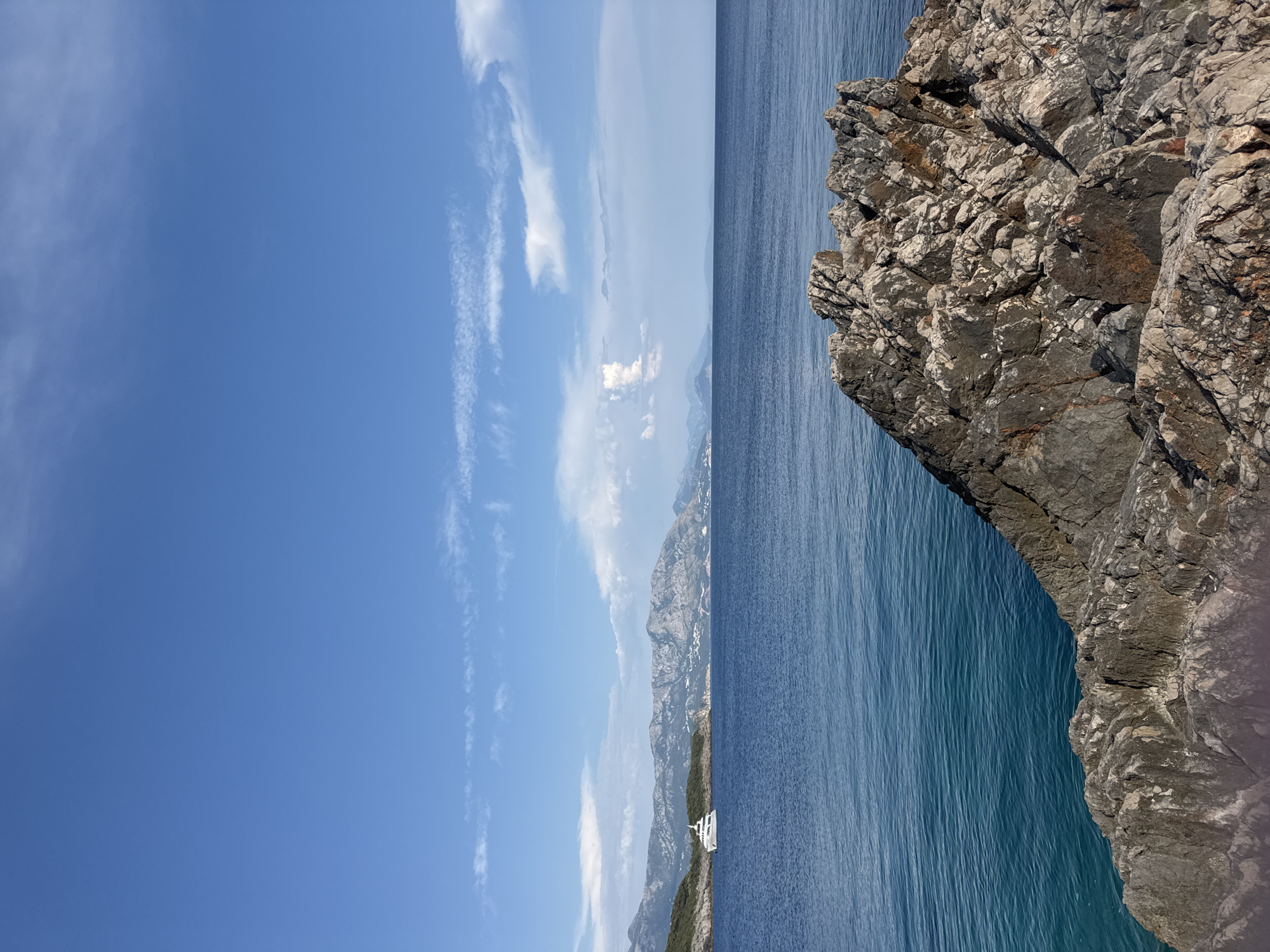 A sunny day unfolds over the coast of Dubraljevina, Montenegro, with a clear blue sky dotted with wispy white clouds. In the foreground, jagged brown rocks dominate the lower right quadrant, their textured surfaces catching the light. Beyond the rocks, the deep blue sea stretches out, meeting a distant, rugged coastline characterized by green hills and mountainous terrain. A white yacht is visible in the distance, anchored near the shore. The water is calm, reflecting the brilliant blue of the sky, and the overall scene conveys a sense of serene coastal beauty.