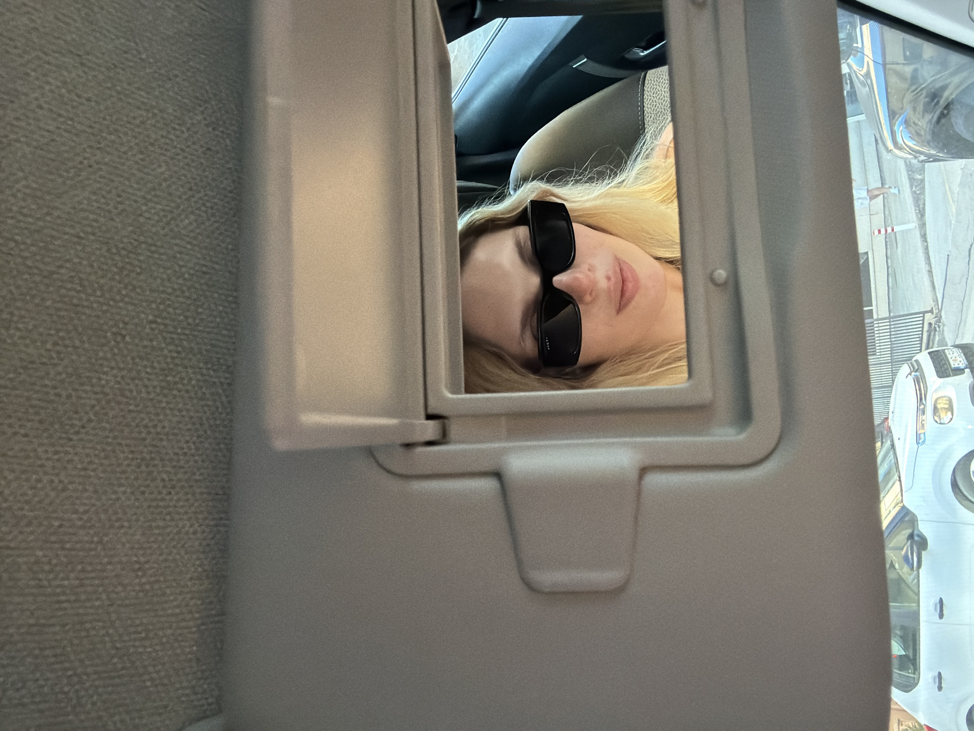 The image captures a casual moment inside a car, with the primary focus on a person reflected in the passenger-side sun visor vanity mirror. The photo itself is rotated 90 degrees counter-clockwise.
In the center of the frame, an open vanity mirror reflects the upper body of a person. The individual has blonde hair and is wearing stylish black rectangular sunglasses, which obscure their eyes. Their face appears relaxed, with lips slightly parted, and their head is tilted to the right.
Around the mirror, the light grey interior of the car's sun visor and ceiling are visible. Through the car windows, particularly to the right of the mirror, a bright outdoor scene is discernible. This includes parts of a white vehicle, possibly a truck or large SUV, and some indistinct background structures suggesting an urban or roadside environment. This exterior view is also partially reflected in the vanity mirror.
The bright natural lighting indicates it is daytime and likely sunny, as further suggested by the use of sunglasses. The activity appears to be a casual selfie or a posed photograph taken using the car's mirror. There is no visible text in the image. The setting is Budva, Montenegro.