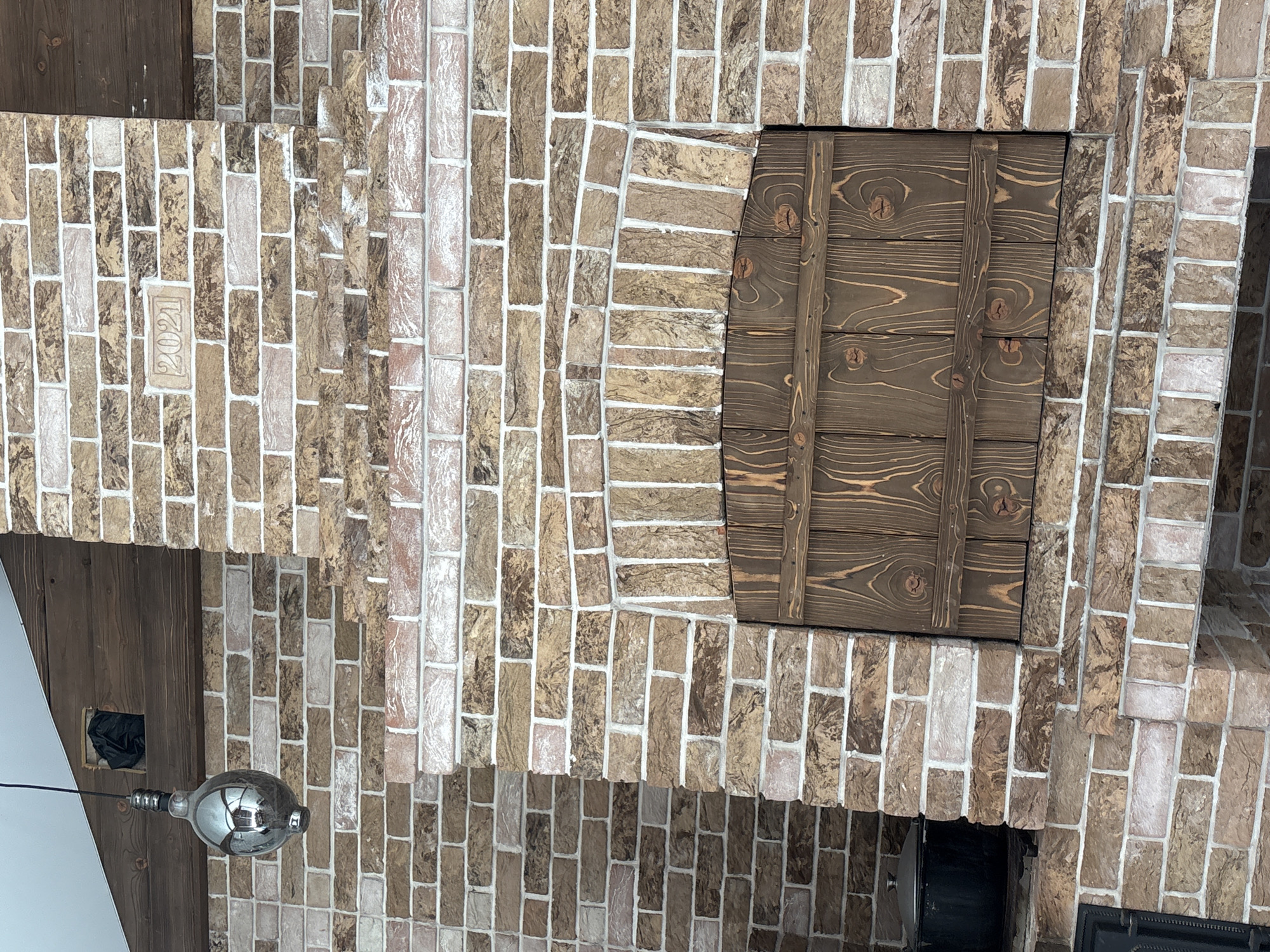 The image shows a brick oven with a wooden door in Georgiyevsk, Russia. The oven is made of light brown and white bricks, and the door is made of dark brown wood with visible grain. Above the oven, there is a wooden beam with a black garbage bag stuffed into a rectangular opening. A silver, bulb-shaped light fixture hangs from a black cord to the left of the oven. A black pot is visible on a shelf to the left of the oven door. The number "2021" is carved into a brick above the oven opening.