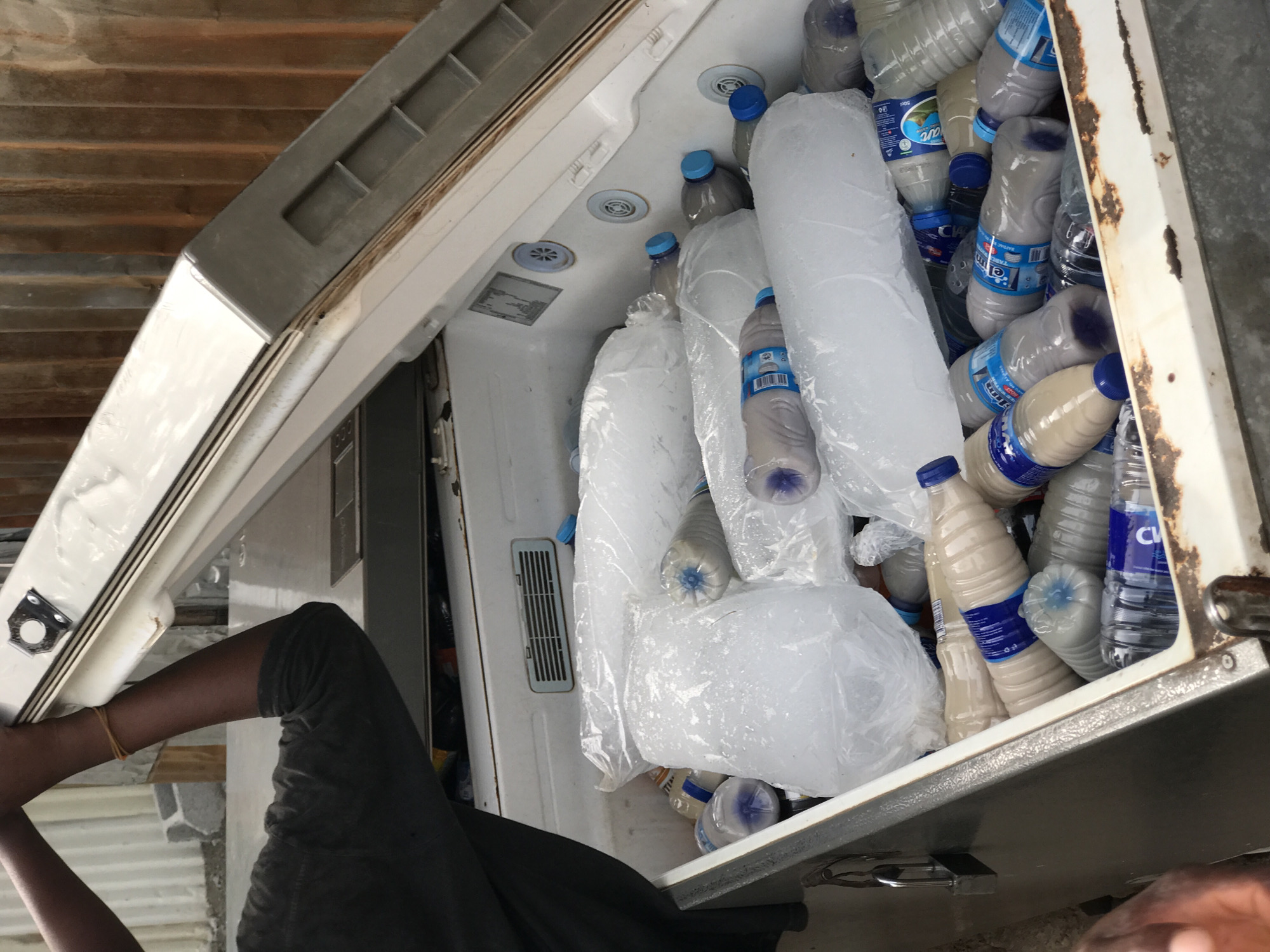 The image shows a downward-angled view into an open, white chest freezer, heavily stocked with plastic bottles and large bags of ice. The freezer appears weathered, with noticeable rust along its top edges, particularly on the right side, suggesting outdoor exposure or age. Inside, numerous clear plastic bottles, many containing water with blue labels (one visible with "50cl" and "pure"), are interspersed with bottles holding a milky-white liquid, also featuring blue labels (some partially legible as "EMIN"). Several large, irregularly shaped blocks of ice, sealed in clear plastic bags, fill the remaining space, indicating a high demand for cooling.
In the lower left, a person's dark-skinned arm and leg are visible, suggesting they are reaching into or standing beside the freezer. A light-colored band is worn on the person's wrist. The background features corrugated metal roofing or siding, indicating an outdoor or semi-outdoor setting, possibly a market stall or roadside vendor. The bright, even lighting points to daytime. The accumulation of ice and cold beverages strongly implies a hot climate, a common characteristic of Abuja, Nigeria. The overall scene depicts a practical setup for selling chilled drinks and ice, essential for refreshment in warm conditions.