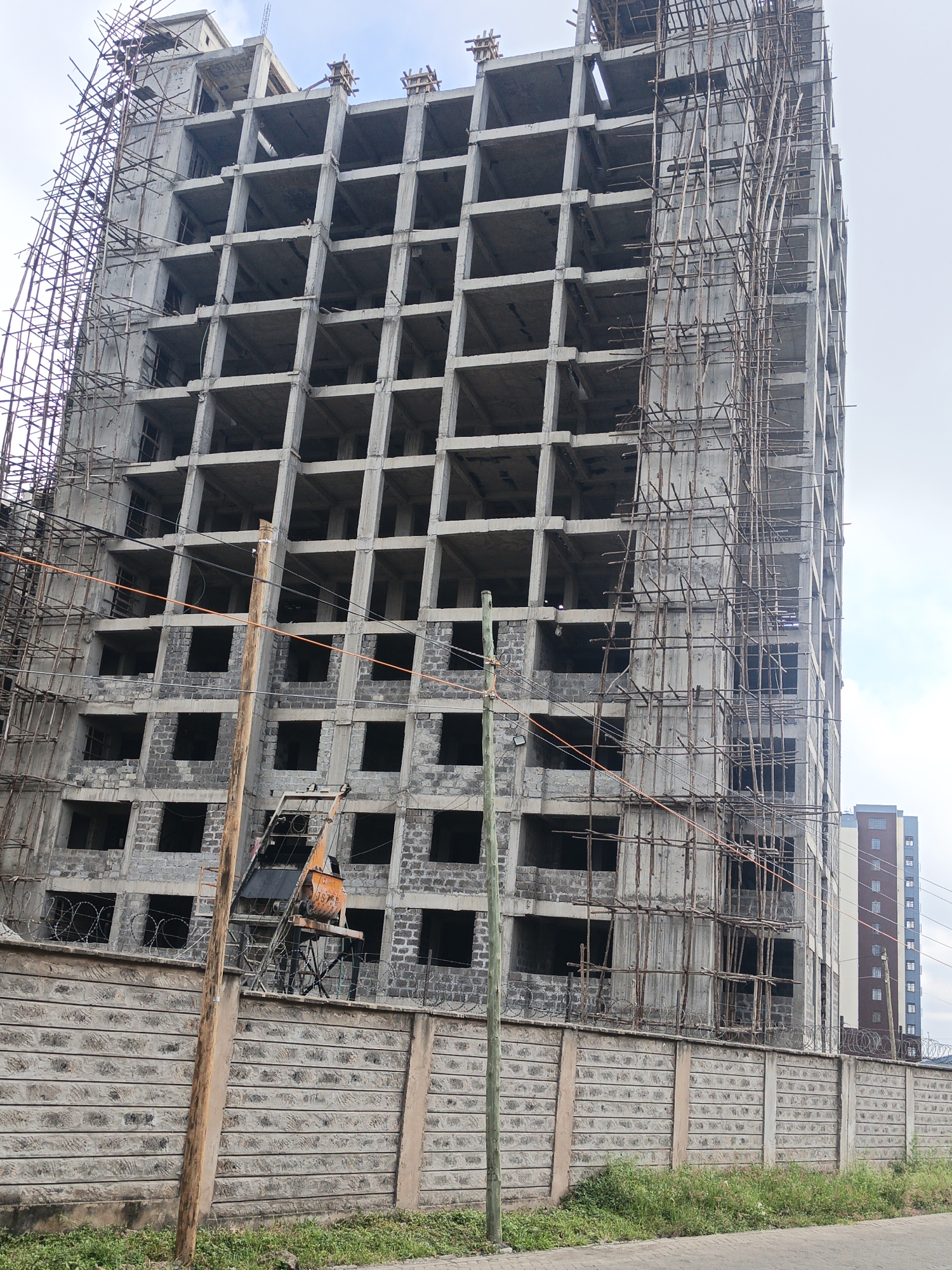 A medium-angle shot depicts an unfinished high-rise building under construction in Nairobi, Kenya. The structure is made of concrete and has a grid-like facade with numerous square openings for windows. The exterior is encased in scaffolding on two sides. At the building's base, a tall concrete wall is topped with barbed wire. An orange cement mixer sits behind the wall. Several wooden utility poles are visible, along with power lines. A completed high-rise building can be seen in the background to the right, suggesting a cityscape. The sky is overcast. The scene appears still, with no visible human activity. The environment consists of unkempt grass and a paved surface.