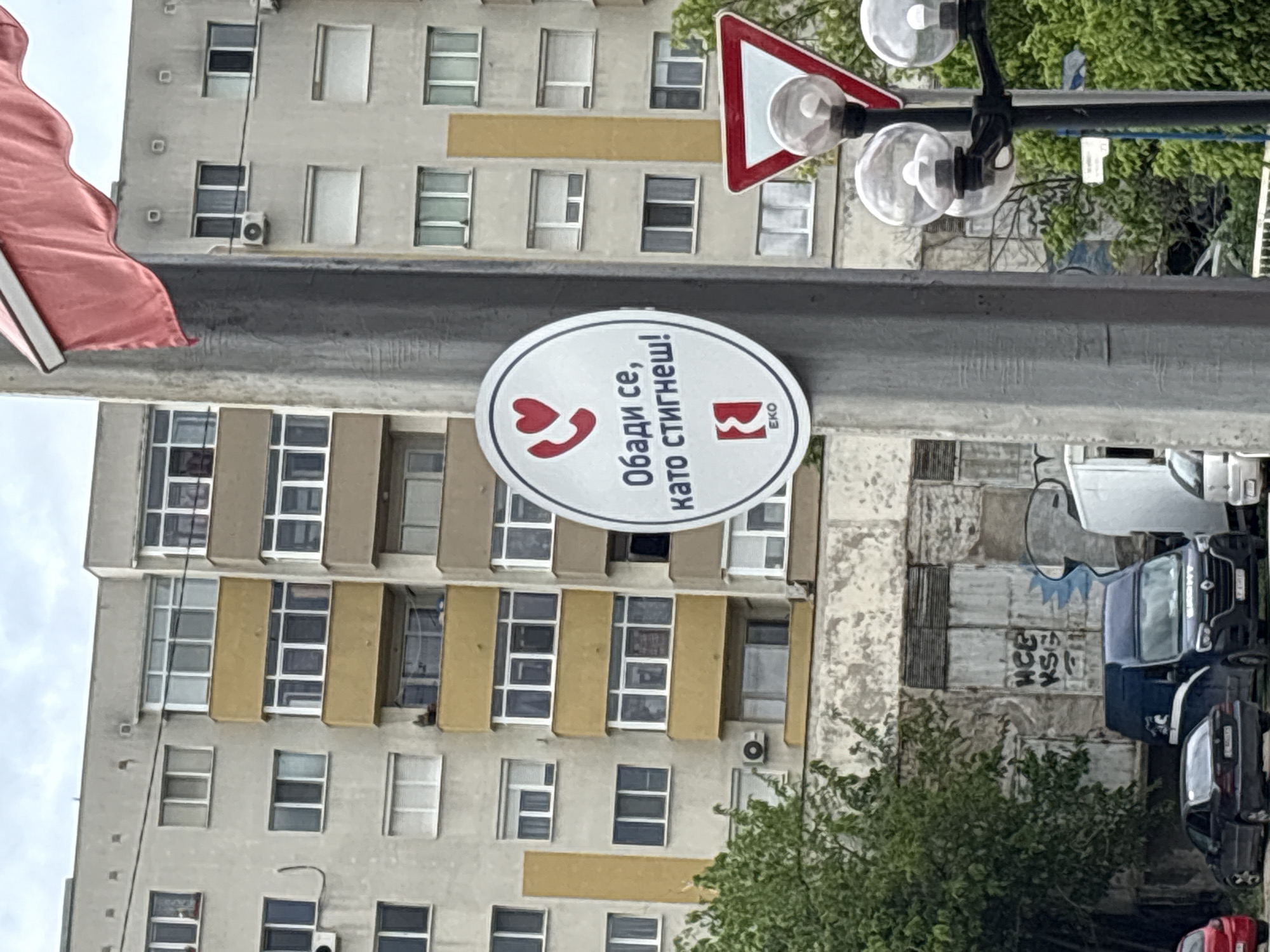 Here is a description of the image:
The image shows a street scene in an urban area. A tall building with multiple windows and yellow-beige architectural details forms the backdrop. A grey metal pole stands in the foreground, and attached to it is a white oval sign with the text "Обади се, като стигнеш!" in Macedonian, along with a stylized phone receiver symbol incorporating a heart and the logo of EKO, a gasoline retailer. Above the sign, there are some streetlights with rounded glass globes and a triangular road sign with a red border. To the right of the pole, there is greenery and several parked cars. The overall environment suggests a street with traffic and commercial activity. The city is presumed to be Unknown, Unknown, which could be the location of the image being taken.