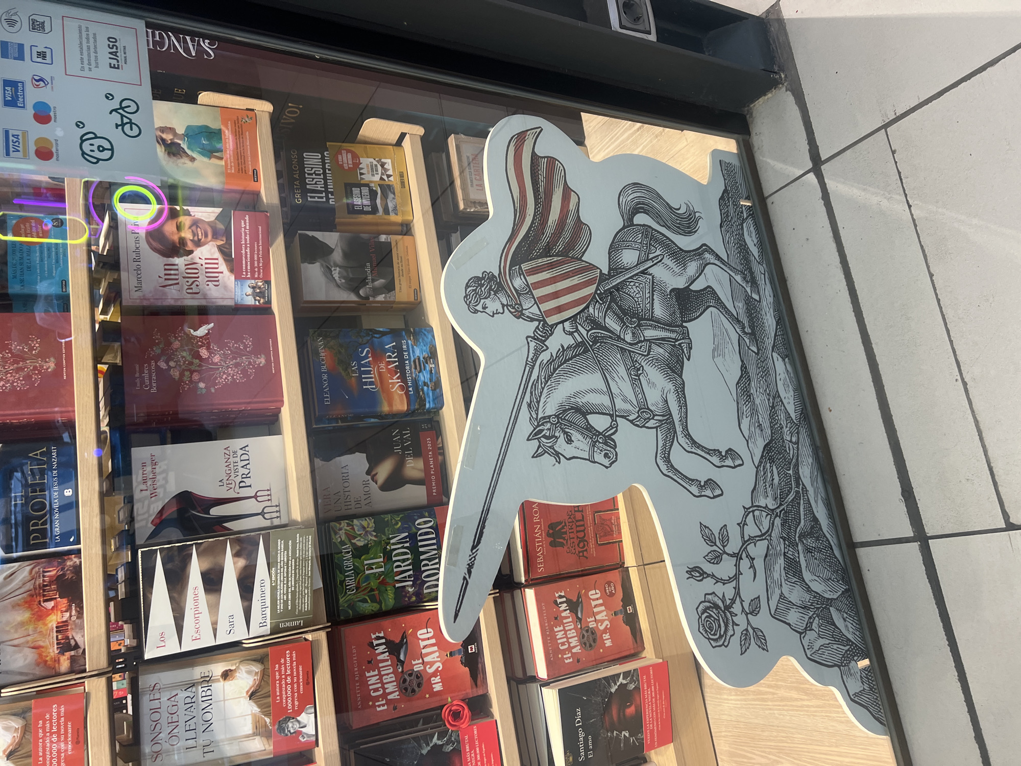 An eye-level shot captures a vibrant bookstore window display, likely in Tarragona, Spain, given the language and context. The scene is brightly lit, suggesting daytime, with sunlight reflecting off the glass. Inside the window, numerous books are neatly arranged on wooden shelves.
A prominent, large cutout figure of a medieval knight on horseback dominates the right side of the display, extending slightly beyond the window frame. The knight, rendered in a monochrome, etched style with a reddish-and-white striped cape and shield, holds a lance. Below the horse, a detailed rose with leaves and rocks are depicted, implying a setting.
Behind and around this cutout, various books are visible. Notable titles and authors include "Aún estoy aquí" by Marcelo Rubens Piva, "EL ASESINO DE INVIERNO" by Greta Alonso, "LA VENGANZA VISTE DE PRADA" by Lauren Weisberger, "Cumbres Borrascosas" by Emily Brontë, "SONSOLES ONEGA LLEVARA TU NOMBRE", "Los Escorpiones" by Sara Barquinero, "EL JARDÍN DORMIDO" by Carla Gracia, "EL CINE AMBULANTE DE MR. SAITO" by Annette Bjergfeldt, "PROFETA", "LAS HIJAS DE SKARA", and "ESTIRPE DE ÁGUILA" by Sebastián Roa. Some books highlight accolades like "PREMIO PLANETA 2025" or "La autora que ha conquistado a más de 1.000.000 de lectores".
In the upper left corner of the window, a white sign displays accepted payment methods (Visa, Mastercard, Maestro, American Express) along with symbols for contactless payment, bicycle parking, and a dog-friendly icon. Below this, a red text box states: "En este establecimiento se denuncian todos los hurtos detectados" (In this establishment, all detected thefts are reported) followed by "EJASO PENAL RETAL", indicating a security measure. The reflection of the street pavement is visible at the bottom right.