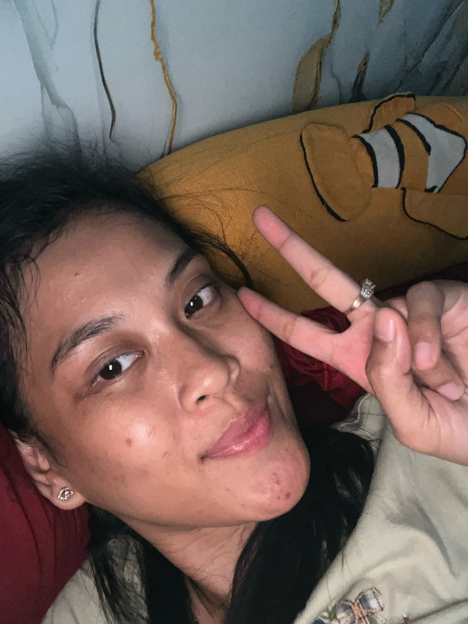 A close-up, selfie-style photograph features a young woman lying down, looking at the camera. She has dark hair, brown eyes, and appears to be wearing makeup. She is making a peace sign with her right hand, which is adorned with a ring. A red or pinkish object, possibly fabric, is visible behind her hand. To her left, a yellow pillow with a cartoon clownfish design is partially visible. The background shows a wall with a marbled or abstract pattern in shades of grey and white, with some brown streaking. The overall lighting suggests an indoor setting, likely a bedroom. The image does not provide any information about the specific location within Kecamatan Pati, Indonesia.