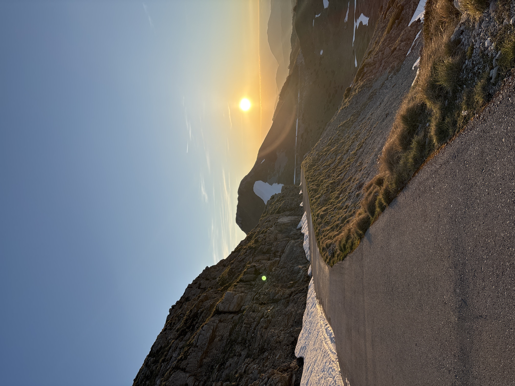 The image shows a mountain road in Pašina voda, Montenegro. The sun is setting behind a mountain range, casting a golden light across the landscape. On the right is an asphalt road and above the road is a slope covered in vegetation with patches of snow. On the left, a steep, rocky mountain face is visible, also with patches of snow. The sky is a clear gradient of blue. There are no people visible in the image.