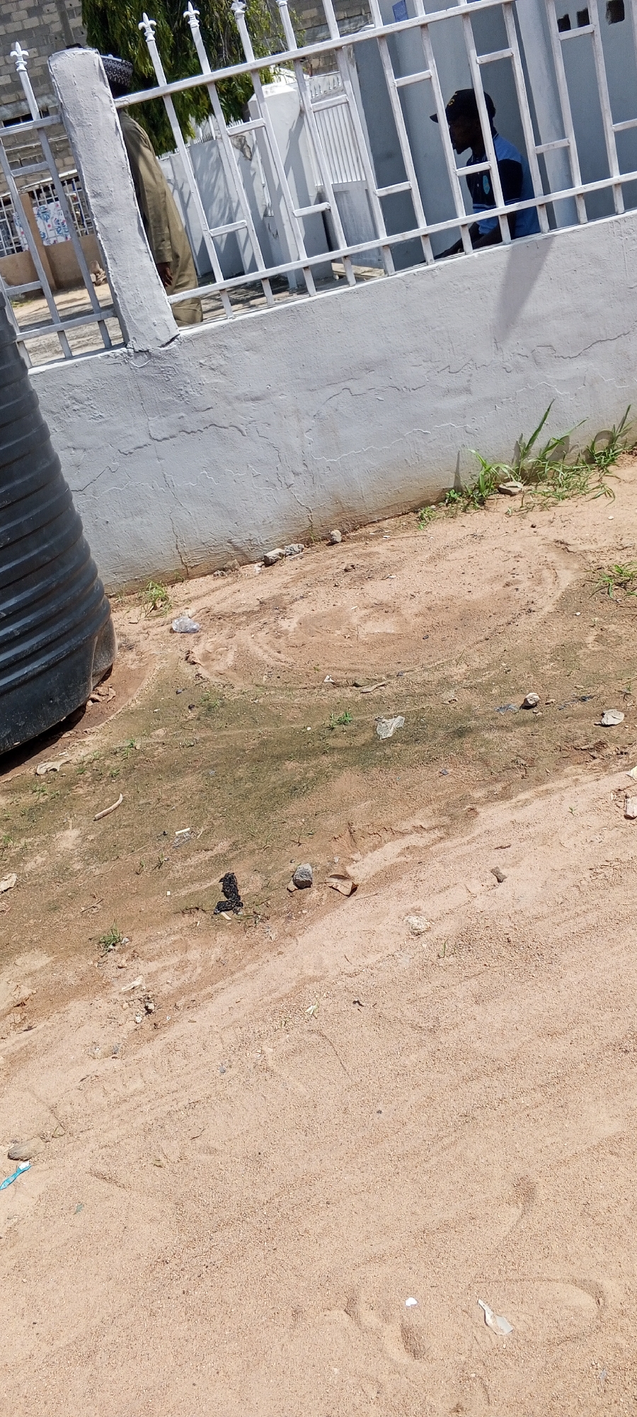 The image depicts an outdoor scene in Jalingo, Nigeria, showing a section of a property with a light-colored concrete wall and a white metal fence. To the left, a portion of a large black cylindrical object, likely a water tank, is visible. Behind the fence, two individuals can be seen. One person, wearing a tan garment and a patterned cap, stands partially obscured by the fence. Another person, wearing a blue and black shirt and a black cap, is seated and looking to the left. The ground in front of the wall is sandy and appears to have some patches of dampness and sparse green vegetation. Scattered on the ground are small rocks and pieces of debris, including a plastic bottle cap. Tire tracks are faintly visible in the sand. The overall impression is of an ordinary residential or communal area.