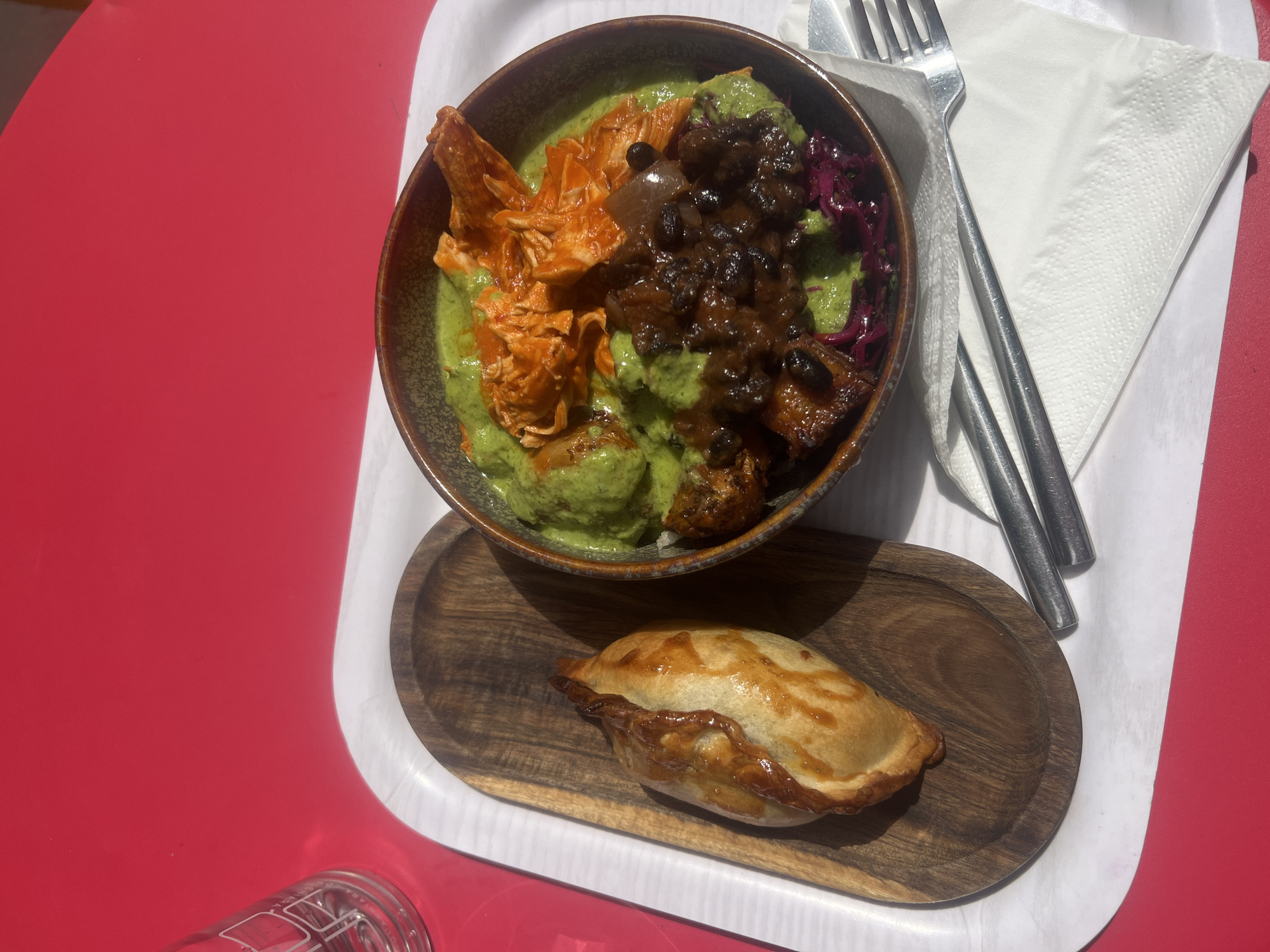 The image depicts a meal served on a white tray, likely in Paris, France, given the style of food. The tray rests on a red surface, possibly a table. A bowl filled with various components dominates the composition: shredded chicken with a reddish-orange sauce, black beans with a dark sauce, shredded purple cabbage, and a green sauce. Adjacent to the bowl is a fork and knife set atop a white napkin. Below the bowl, on a small wooden platter, sits a golden-brown empanada-like pastry. A clear glass, partially visible in the lower left corner, suggests a beverage was also served. The overall lighting is bright, indicating daytime.