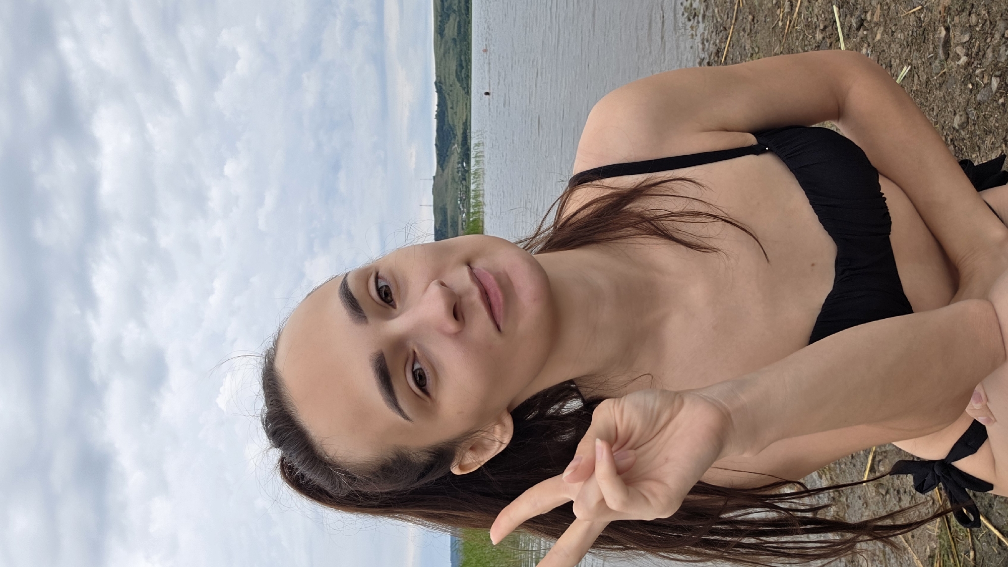 A young woman with dark hair pulled into a ponytail is shown from the chest up, posing for a selfie outdoors. She is wearing a black bikini. She is holding up two fingers in a peace sign with her left hand. She is smiling slightly and looking at the camera. The background shows a body of water, with reeds and hills in the distance. The sky is overcast. The location is the shore of the reservoir in the village of Kupoyarovo, Russia.