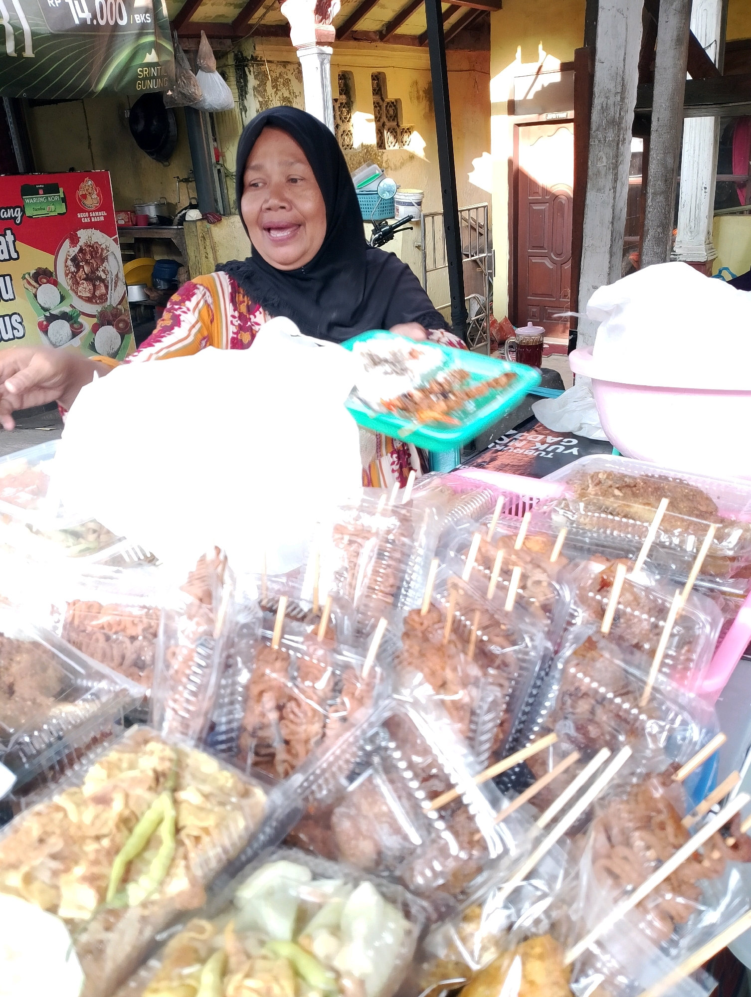 A woman wearing a black hijab and a patterned shirt is standing behind a food stall in Probolinggo, Indonesia. She is holding a green tray with food and smiling at the camera. In front of her, there are several plastic containers filled with food items, many of which are on skewers. Some of the food appears to be grilled meat, while other items are less discernible. Behind the woman, there are more food items, cooking equipment, and a doorway. The stall is located outdoors, with a yellow wall and wooden pillars visible. The overall impression is that of a bustling street food vendor scene.
