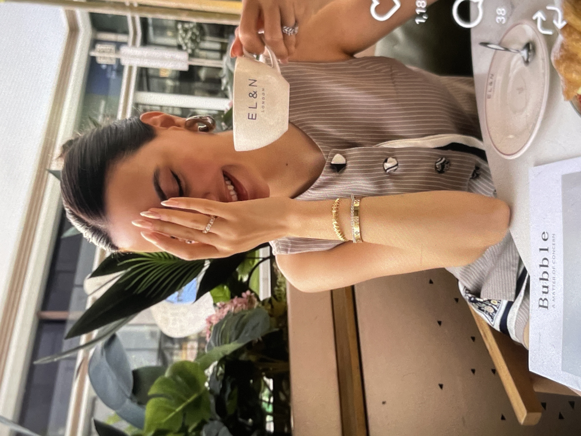 A woman with dark hair pulled back is sitting at a table and holding a coffee cup with the logo "EL & N LONDON" on it. She is wearing a grey and white striped sleeveless top and several pieces of jewelry, including rings and bracelets. She is smiling and covering part of her face with her hand. In front of her is a small plate with a spoon on it and a croissant. The background appears to be an indoor cafe with plants and blurred out patrons. The image also contains superimposed social media icons and text, including a heart icon with "1,18" next to it, a speech bubble icon with "38" next to it, and text that reads "Bubble A MATTER OF CONCERN".