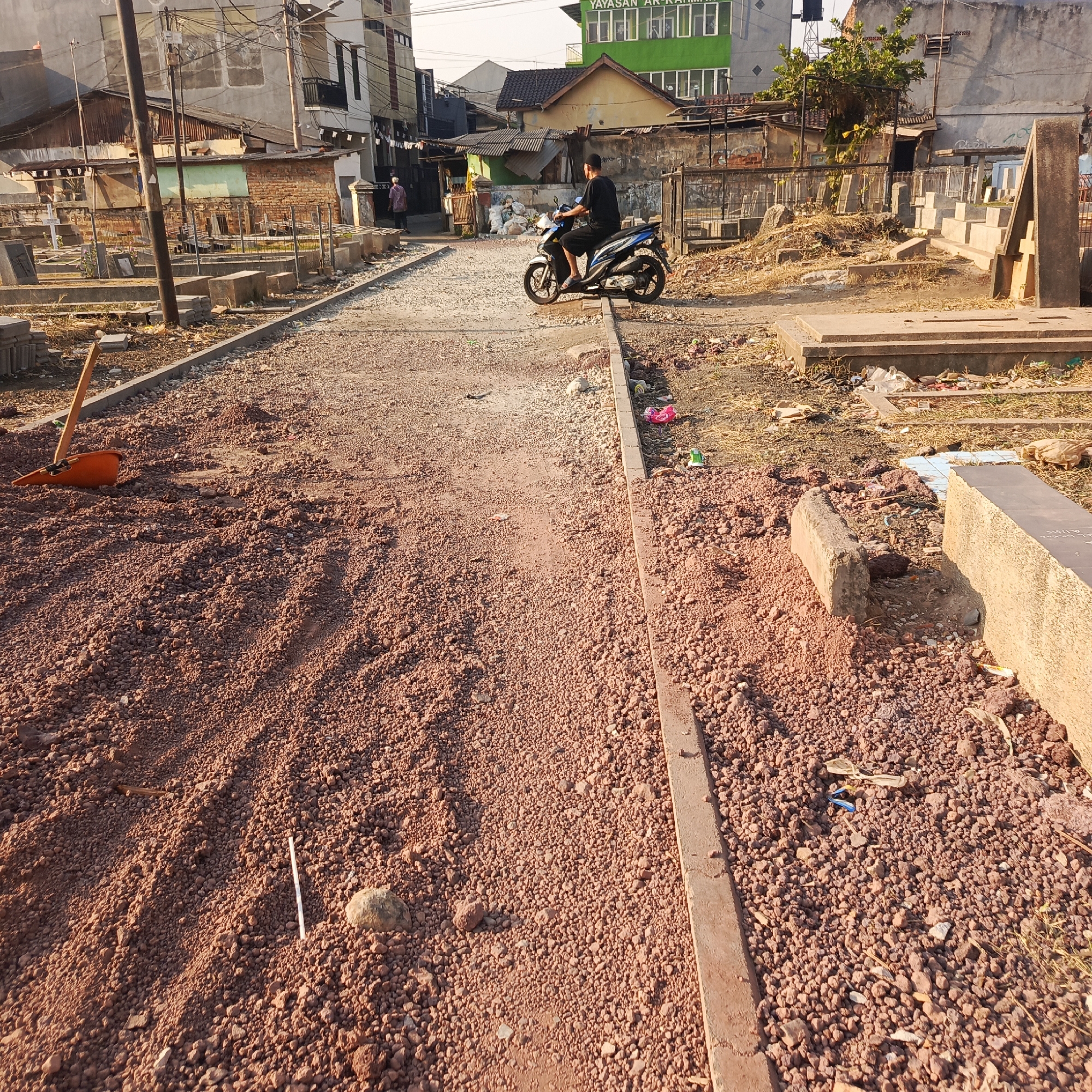 The image depicts an outdoor scene, likely a cemetery or a burial ground undergoing renovation or construction in South Jakarta, Indonesia. The foreground is dominated by loose reddish-brown gravel and soil, suggesting fresh excavation or landscaping. A wooden-handled shovel with an orange scoop lies embedded in the gravel on the left. A thin white stick and a small rock are also visible in the gravel. A distinct border, possibly a curb, runs along the right side of the gravel path.
In the midground, a person is riding a blue and white motorcycle away from the camera. The motorcycle is positioned on a path covered with lighter-colored gravel. Adjacent to this path, to the left and right, are rows of tombstones and grave markers, some appearing aged and weathered, others more modern and concrete. Some graves are surrounded by low concrete borders. The environment appears somewhat unkempt, with dry grass and scattered debris, including plastic bags and wrappers, around the graves.
The background reveals a mix of buildings. To the left, there are multi-story buildings with a somewhat worn appearance, electrical wires, and poles. A smaller, dilapidated structure with a corrugated metal roof and brick walls is also present. In the center background, a bright green building with multiple windows stands out, labeled "YAYASAN AR-RAHMAN". Further in the background, more residential or commercial buildings are visible, some with visible damage or age. The sky is clear and bright, indicating daylight, possibly late afternoon given the warm, low-angled light casting shadows. The overall impression is of a working or transitional urban space, juxtaposing the final resting place with ongoing activity and development.